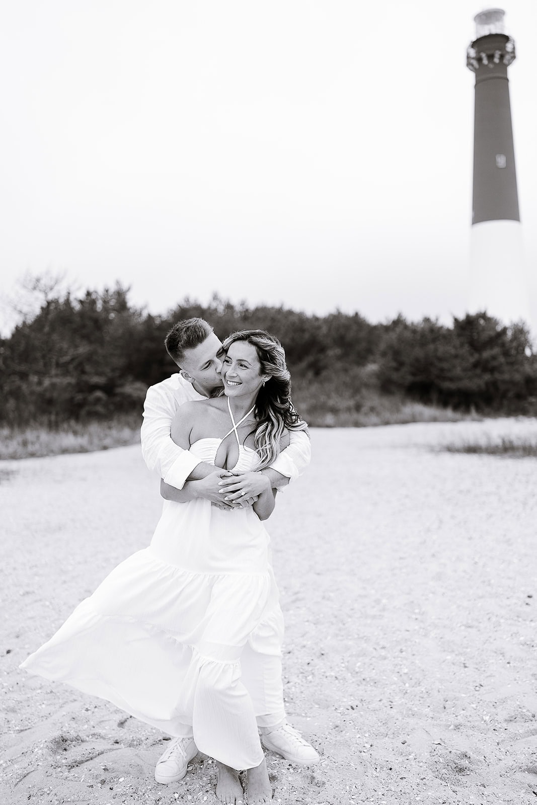Couple embracing on sandy beach with Barnegat Light lighthouse in the background, captured in black and white.