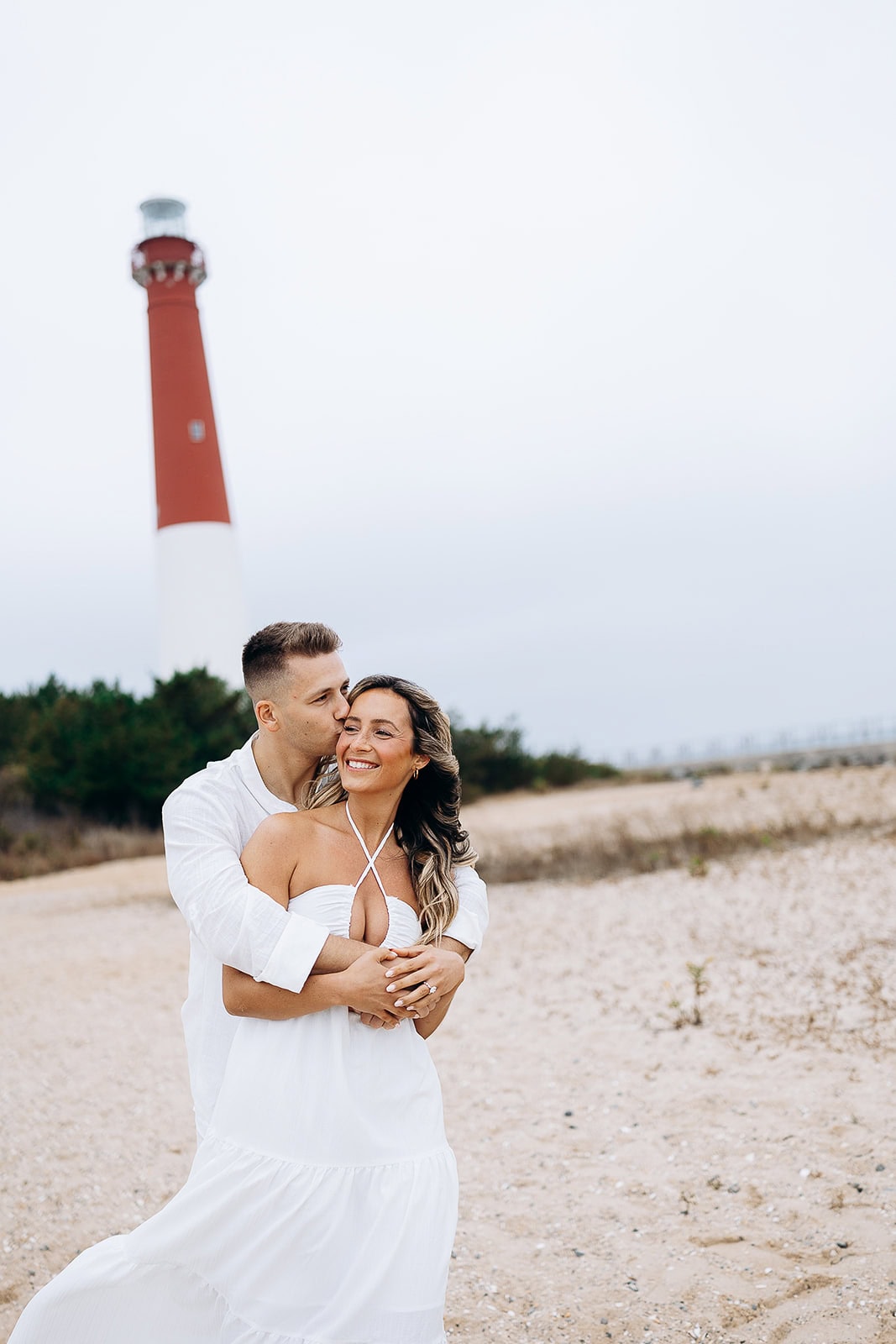 Couple in white clothing embracing on a sandy beach with Barnegat Light Lighthouse in the background under an overcast sky.