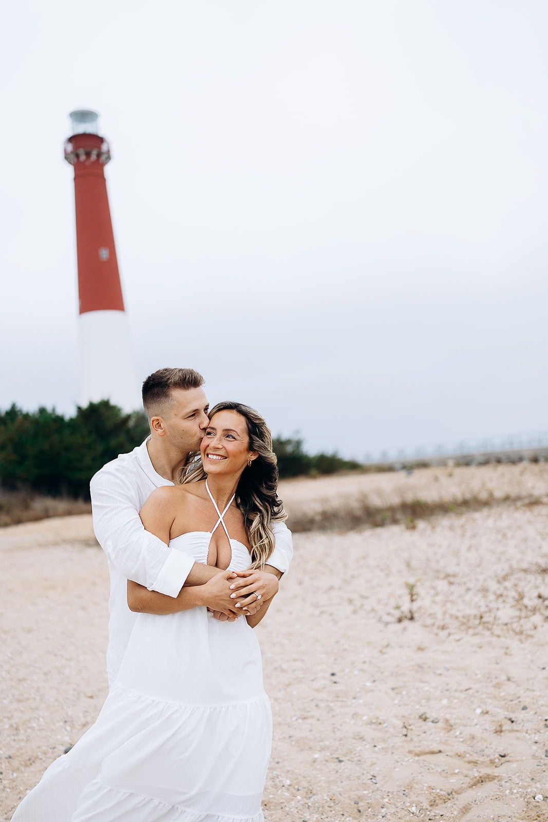 Couple in white attire embracing on sandy beach with Barnegat Light Lighthouse in the background under overcast sky.