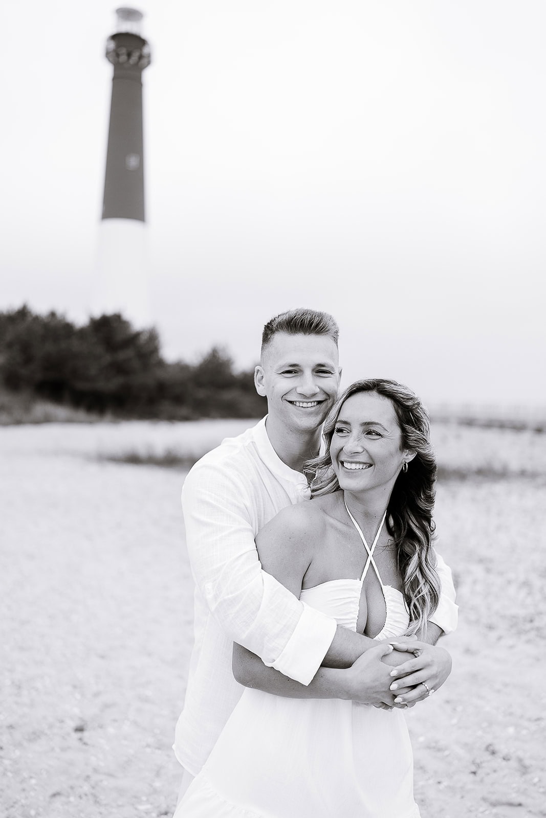 Black and white image of a couple embracing on a sandy beach with Barnegat Light lighthouse blurred in the background.