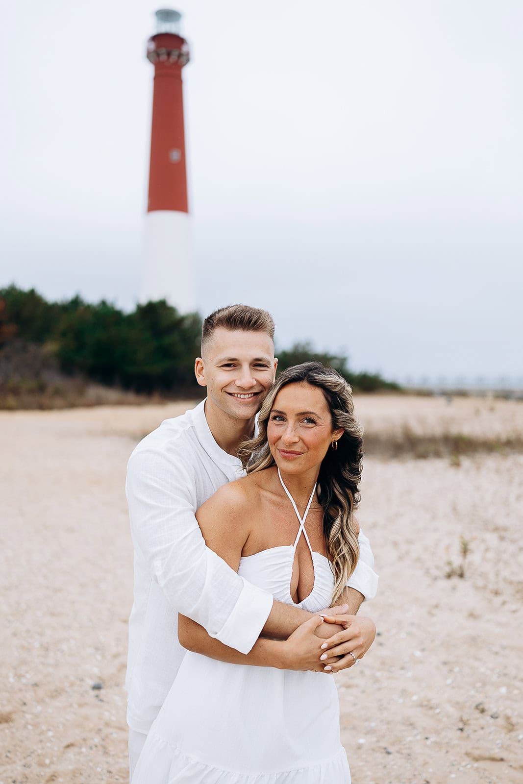 Couple dressed in white embracing on a sandy beach with Barnegat Light lighthouse in the background under overcast sky.