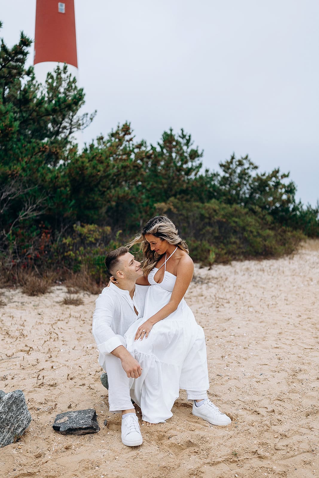 Couple dressed in white seated on sandy beach near green shrubs with red and white Barnegat Light lighthouse in background.