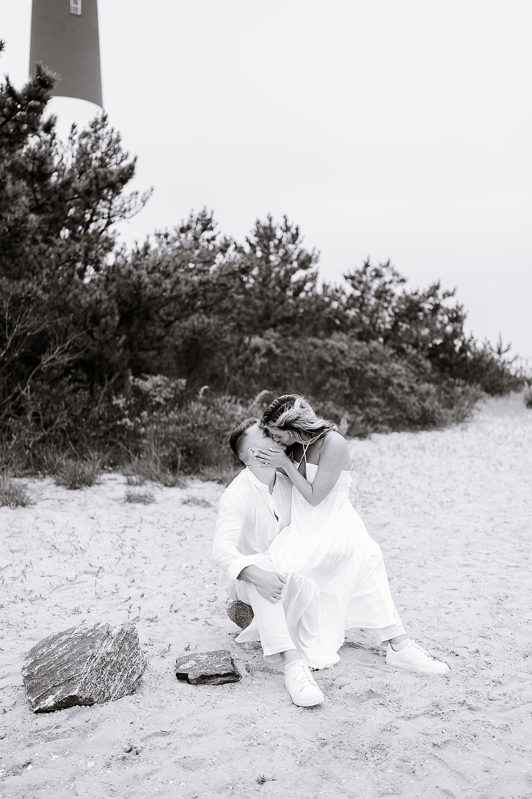 Couple in white clothing sharing a kiss while seated on sand near Barnegat Light lighthouse with dense greenery in the background.