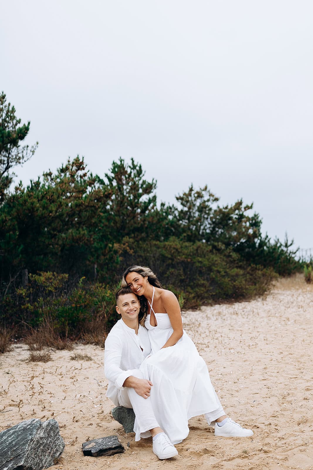Couple dressed in white sitting on sandy dunes near green shrubs under an overcast sky.