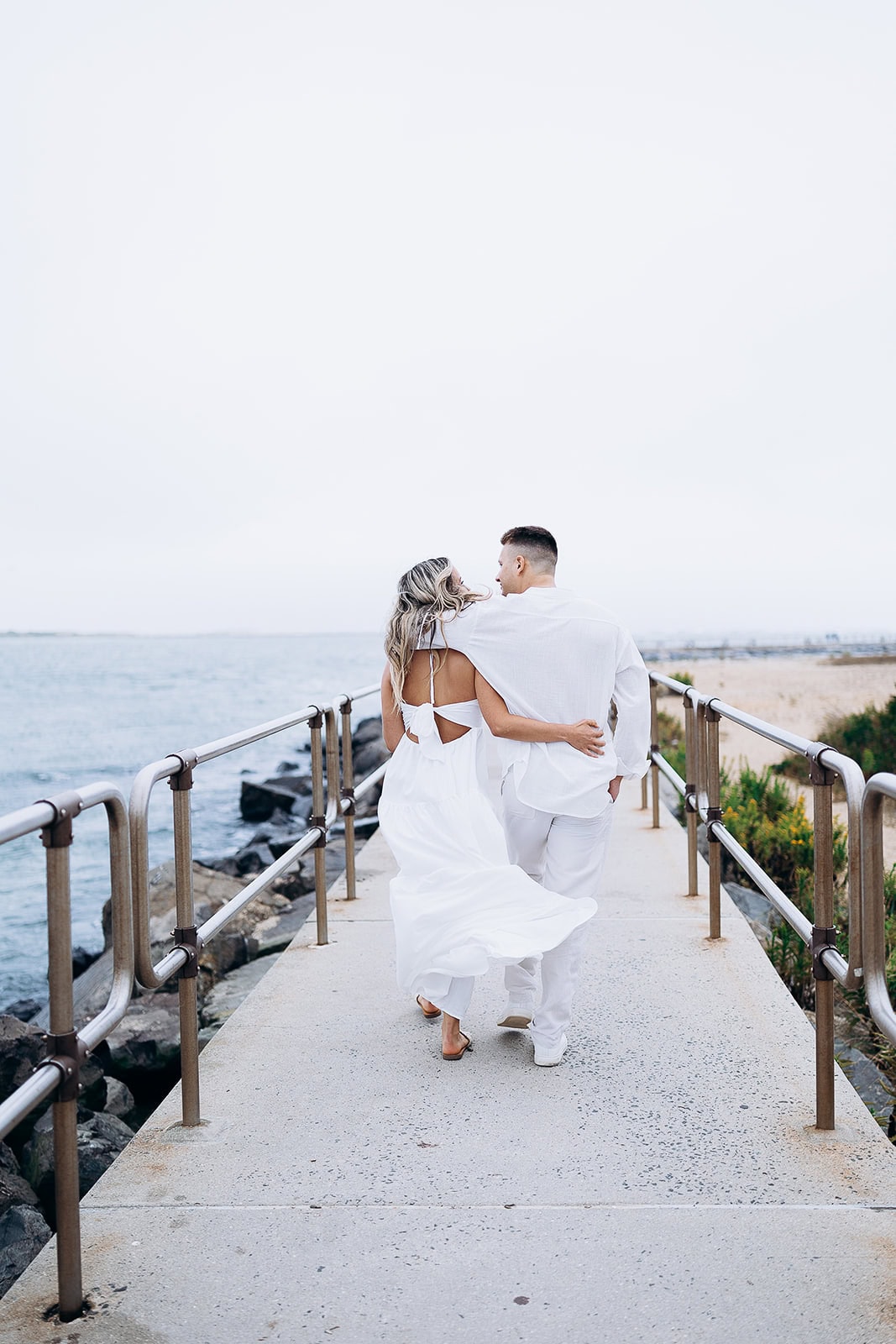 Couple walking arm in arm on a concrete pier with metal railings beside calm ocean waters and sandy shore in Barnegat Light.