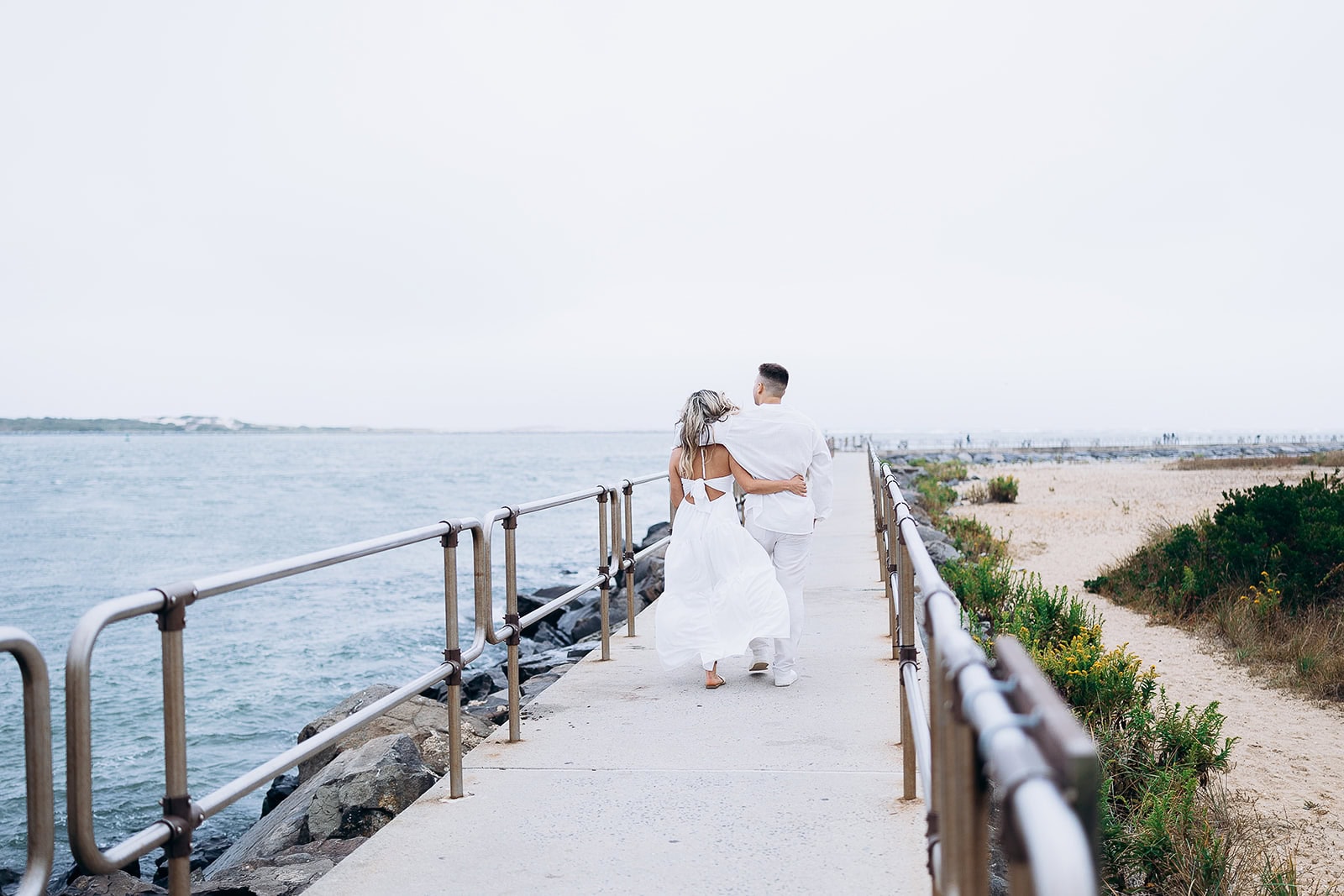 Couple in white clothing walking arm in arm down a concrete pier with metal railings beside the ocean at Barnegat Light, New Jersey.