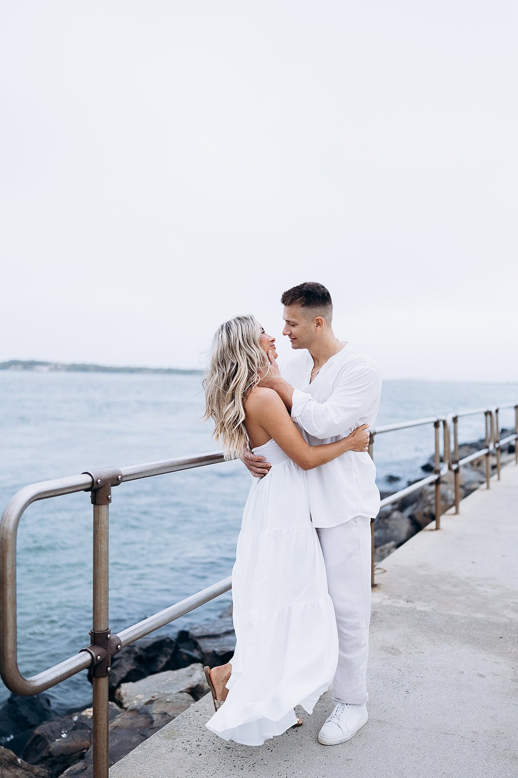 Couple dressed in white embracing on a concrete pier with metal railing by the water in Barnegat Light, NJ.