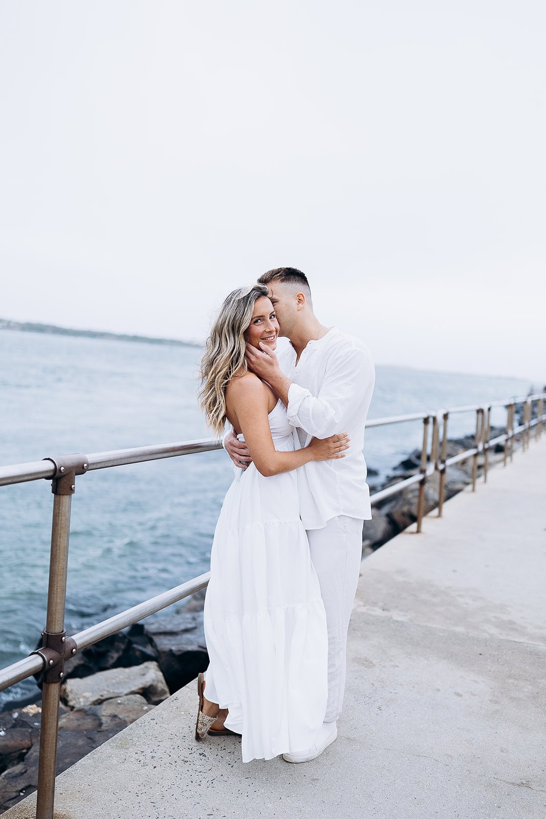 Couple in white outfits embracing on a concrete pier with metal railing beside the ocean at Barnegat Light, New Jersey.