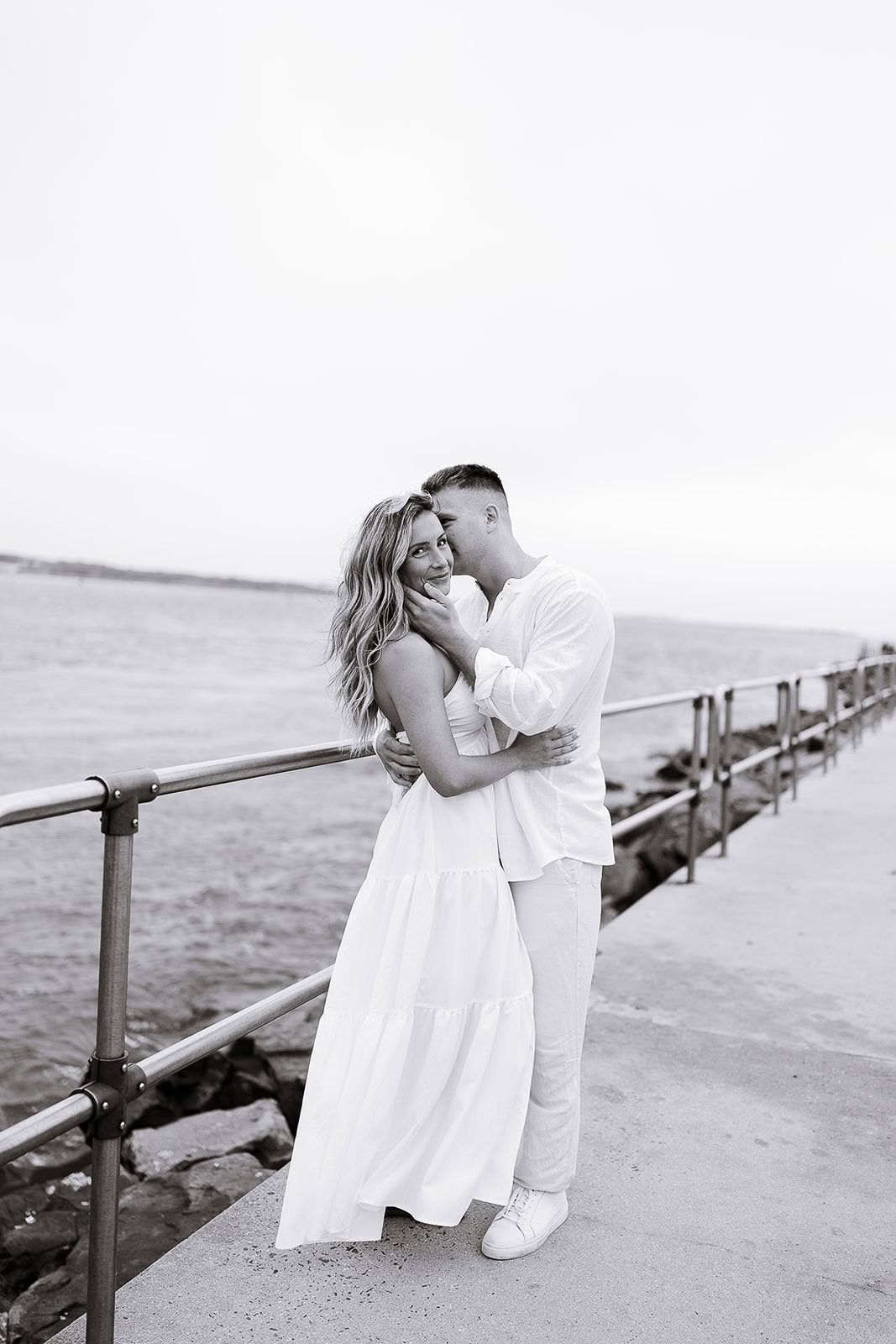 Black and white photo of a couple embracing on a concrete pier with metal railings by the water in Barnegat Light, NJ.
