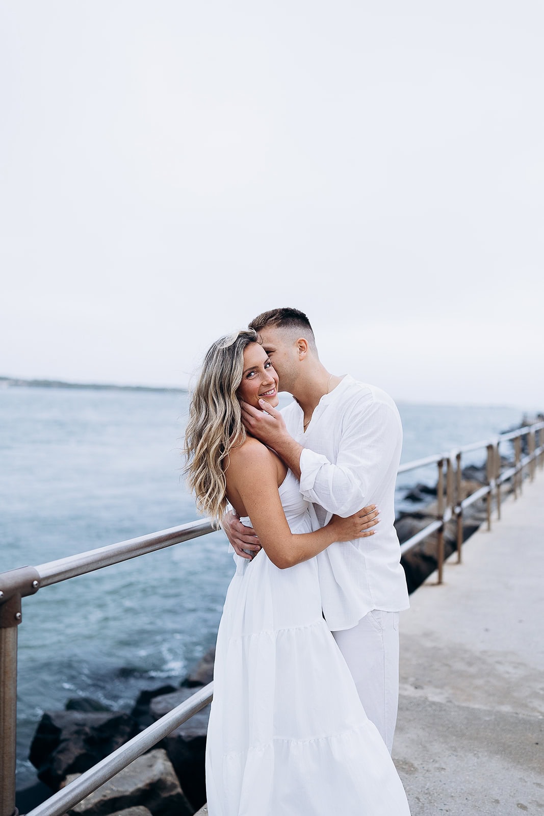 Couple dressed in white embracing by the waterfront on a concrete pier with metal railings and calm sea in Barnegat Light, NJ.