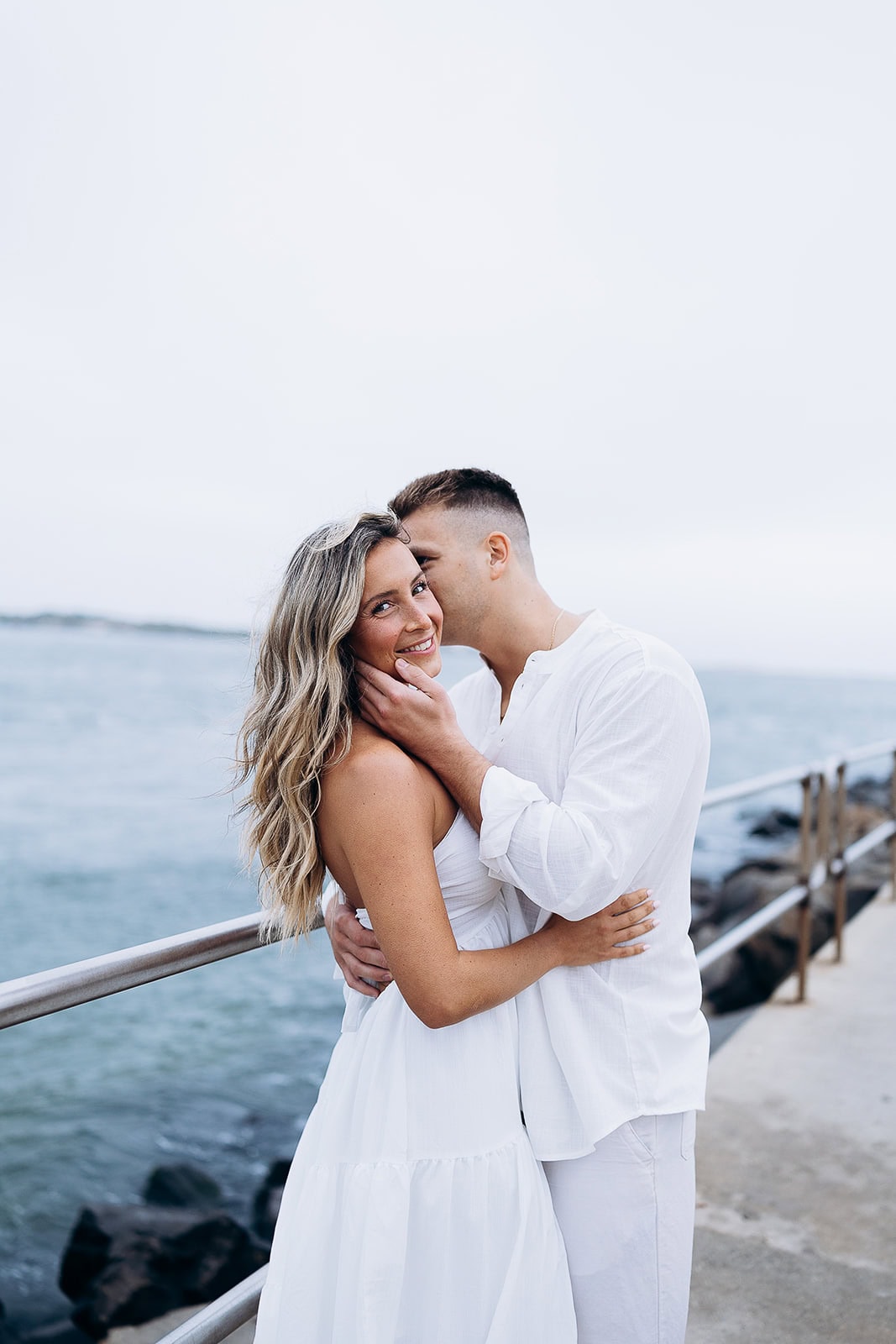 Couple embracing on a waterfront pier in Barnegat Light, the man gently holding the woman's face as she smiles, both dressed in white with ocean and rocks in the background.