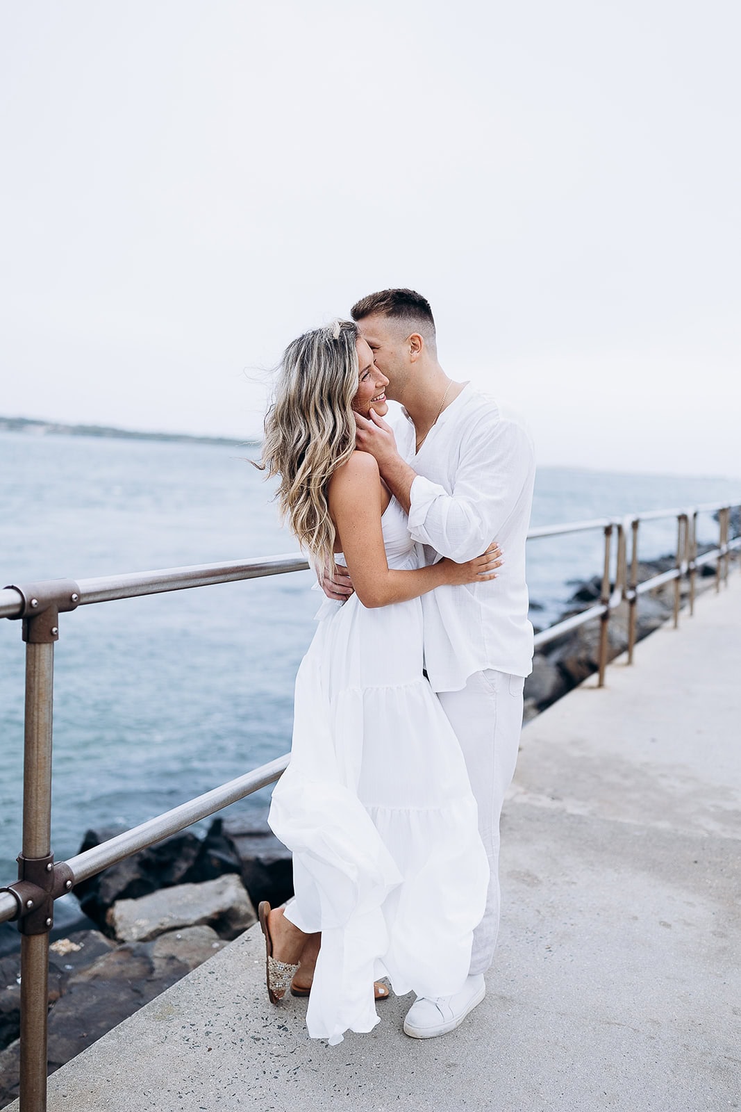 Couple embracing on a concrete pier by the water, dressed in flowing white garments, with a metal railing and rocky shoreline in the background.