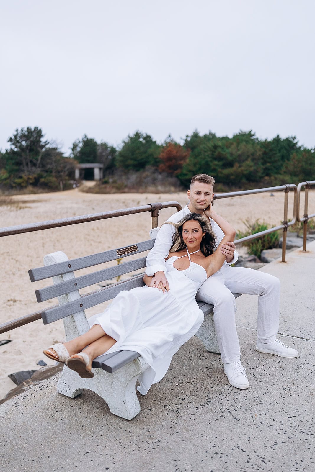Couple dressed in white seated on a gray bench by the beach with greenery in the background at Barnegat Light, NJ.