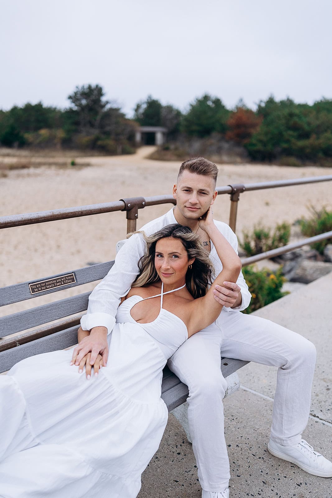 Couple dressed in white sitting on a bench near a sandy beach with greenery in the background at Barnegat Light, NJ.
