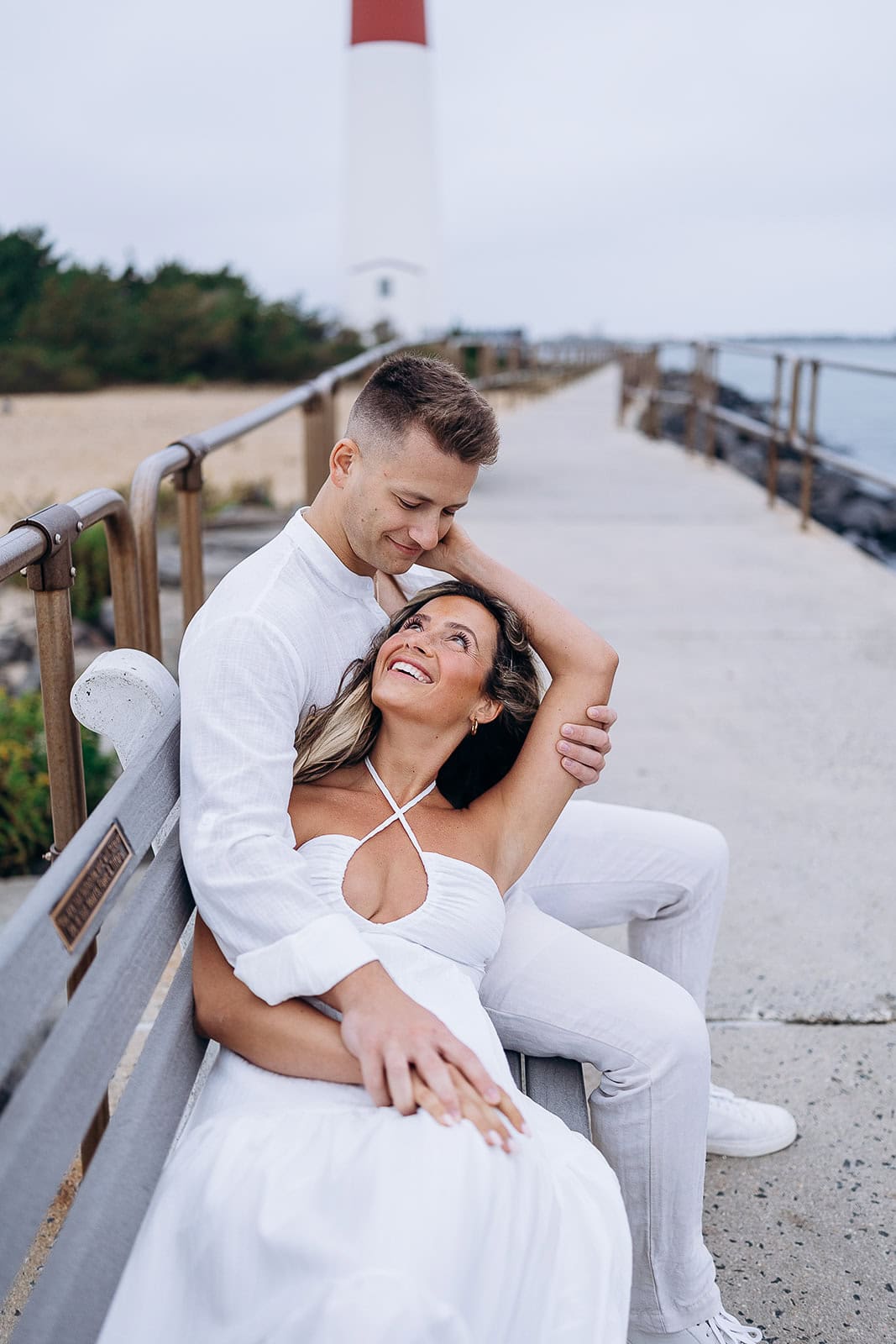 Couple dressed in white seated on a bench at Barnegat Light pier, with a lighthouse and ocean in the background.