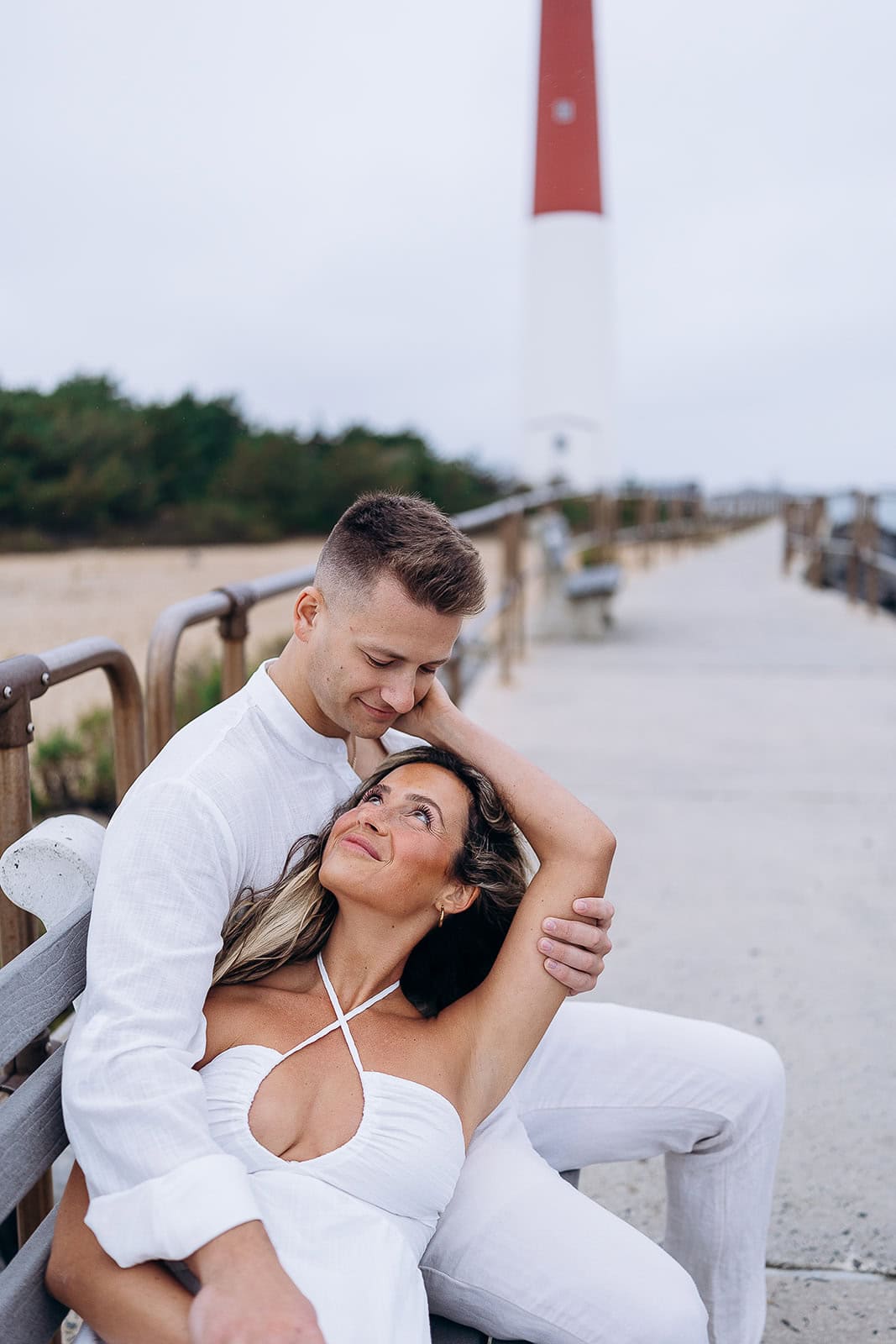 Couple dressed in white seated on a bench by a pier with Barnegat Light lighthouse in the background, Barnegat Light, NJ.