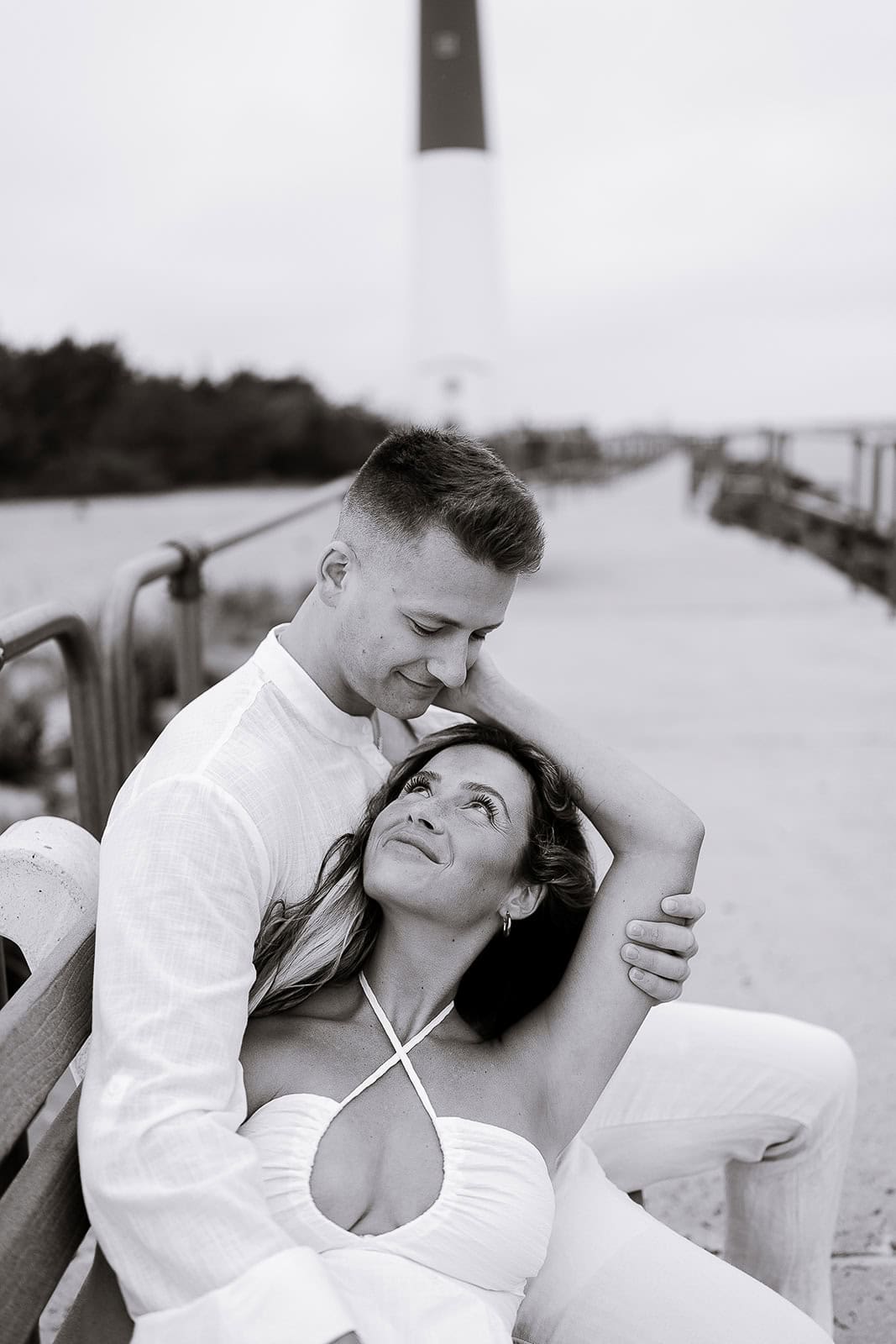 Black and white photo of a couple seated on a bench near Barnegat Light lighthouse, the man gently embracing the woman who looks up at him affectionately.
