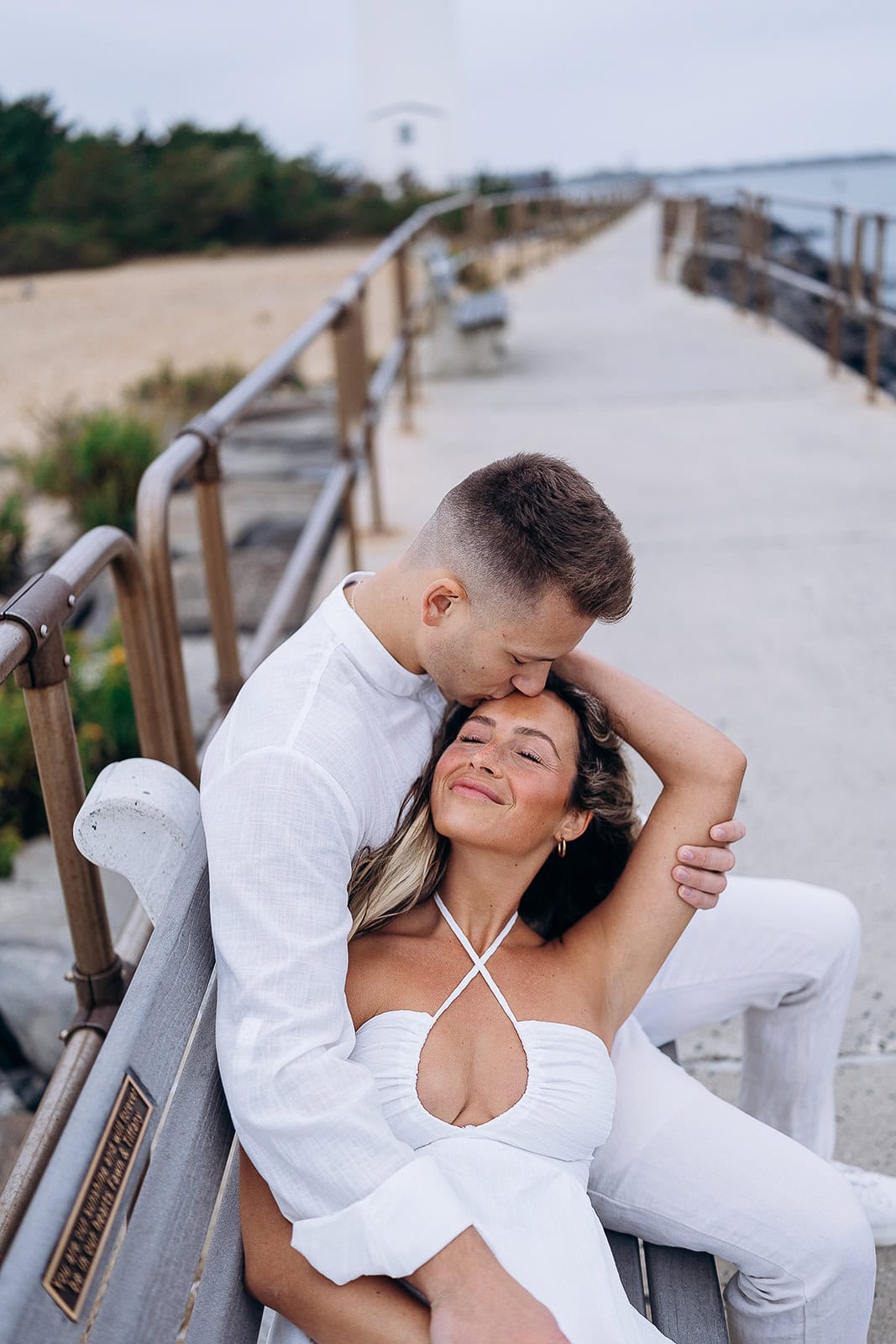 Couple in white outfits seated on a wooden bench by a coastal walkway at Barnegat Light, the man kissing the woman's forehead in an intimate embrace.