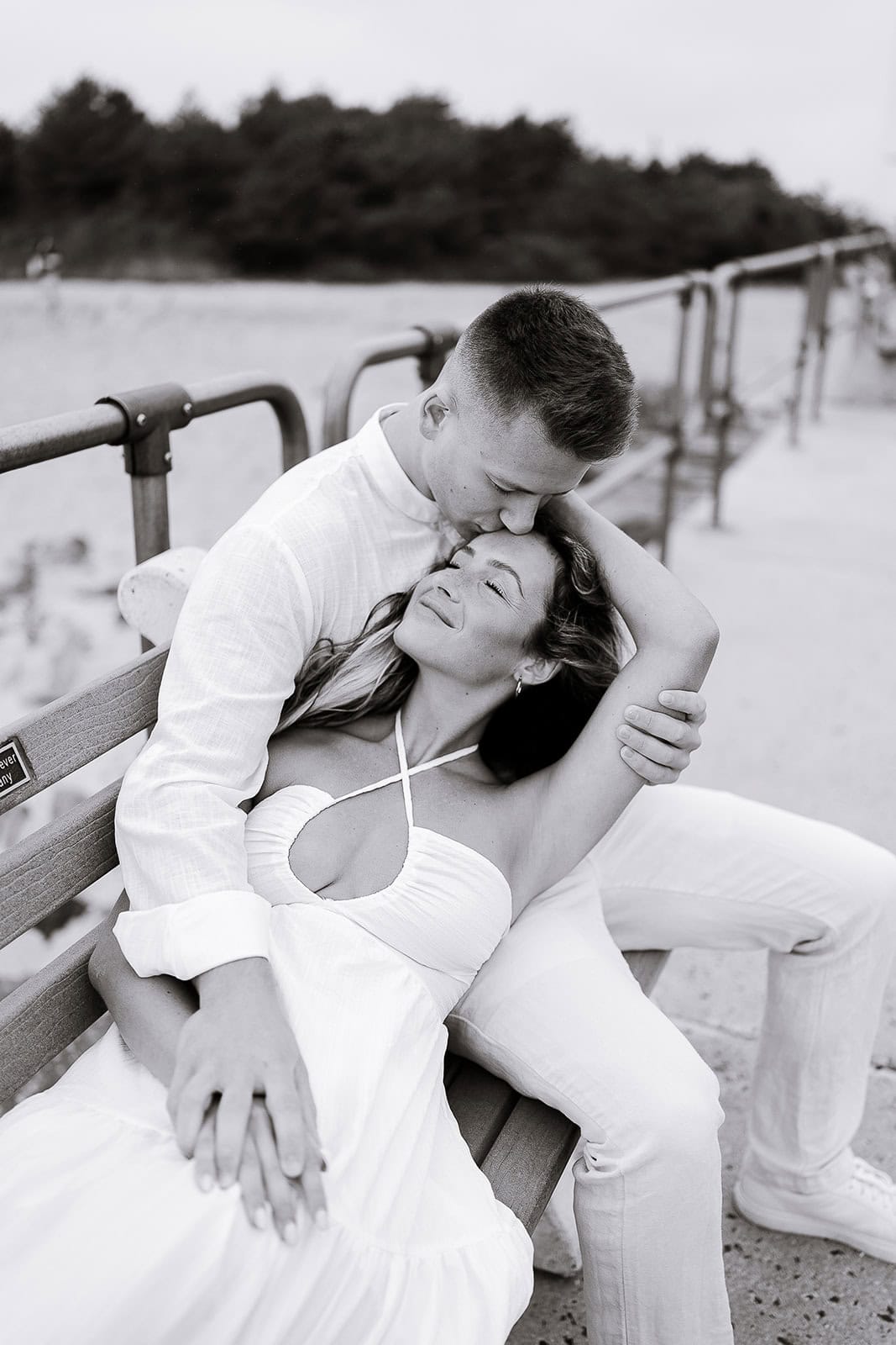 Couple seated on a bench by the beach, the man gently kissing the woman's forehead as she leans back with eyes closed, showcasing a serene and intimate moment.