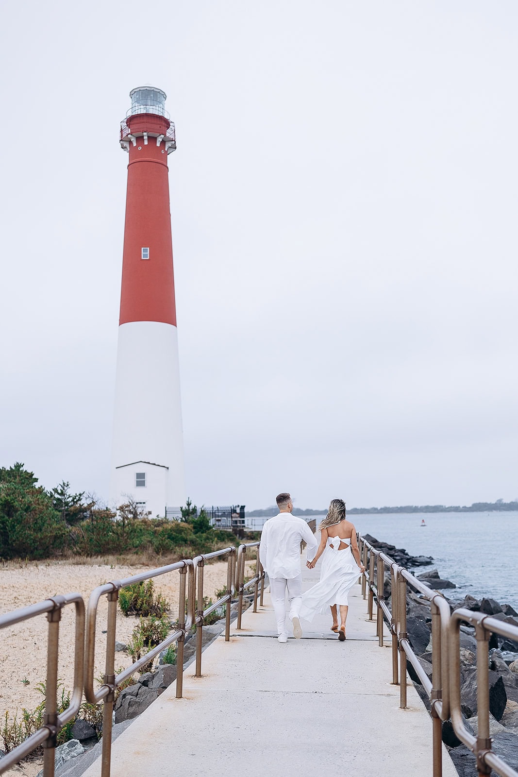Couple walking hand-in-hand on a pier near Barnegat Light lighthouse in New Jersey, overcast sky and calm water in background.