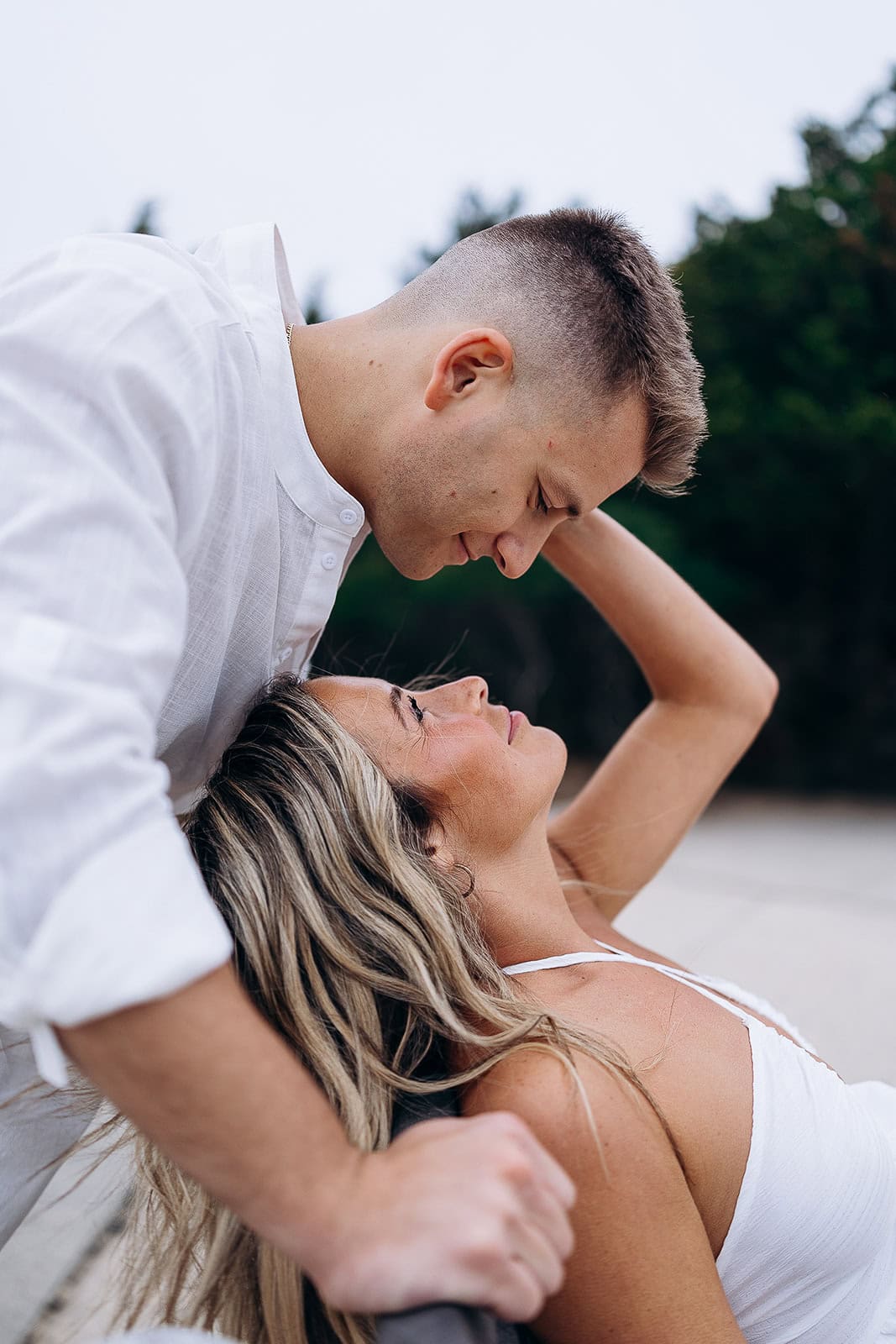 Close-up of a man in a white shirt leaning over a woman in a white dress, both gazing tenderly at each other outdoors with blurred greenery in the background.