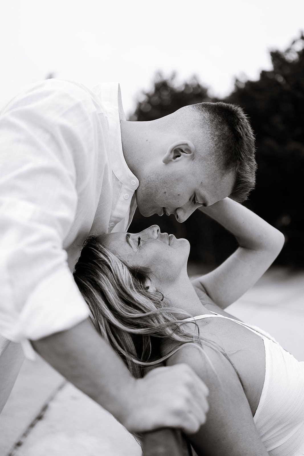 Black and white close-up of a man leaning over a woman, their faces nearly touching, with a blurred natural background.