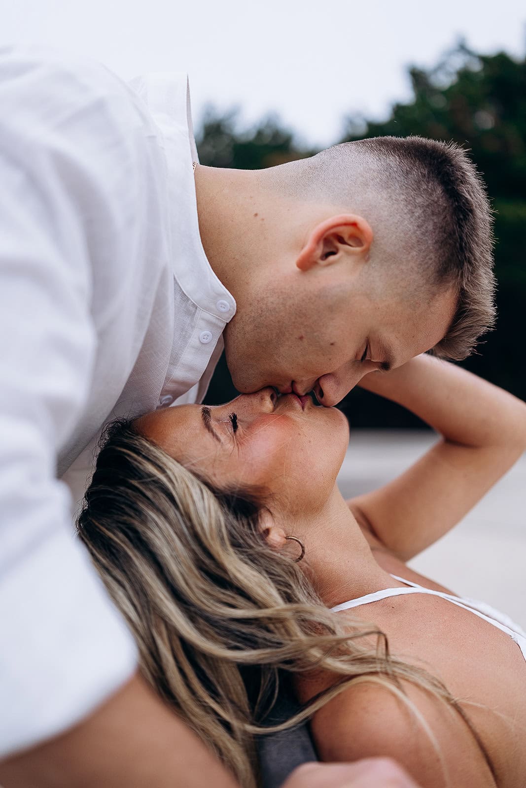 Close-up of a couple sharing a gentle kiss, the man leaning over the woman with eyes closed, set against a blurred natural background.