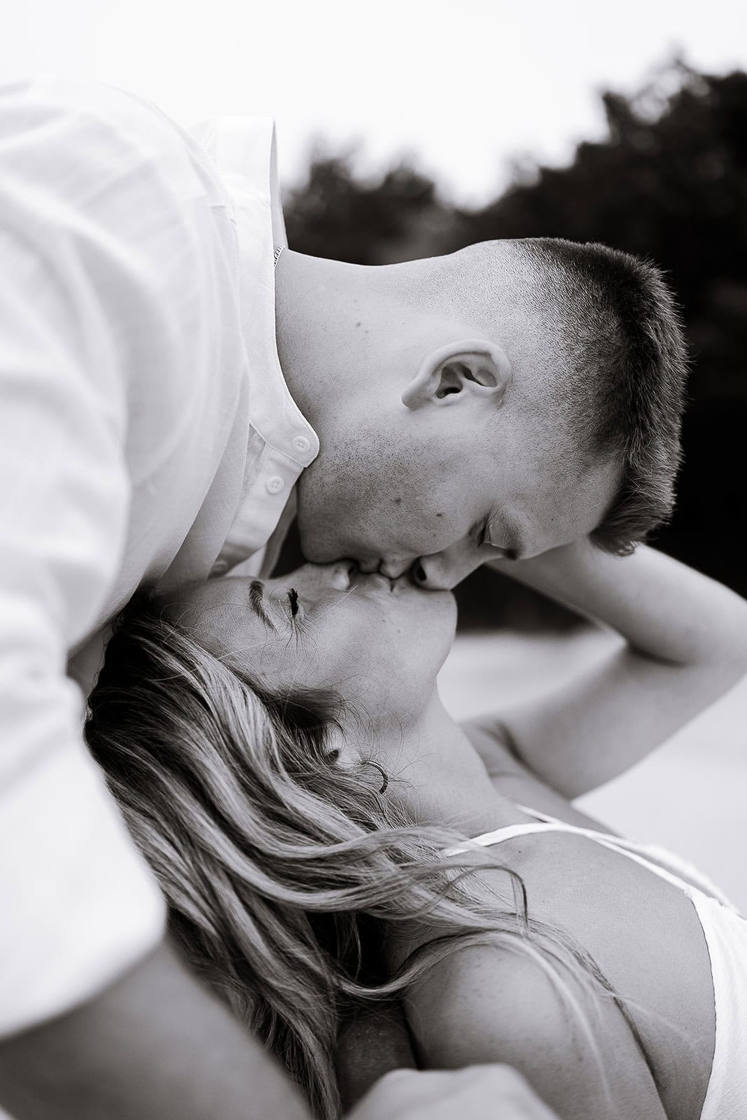 Black and white close-up of a couple sharing a tender kiss, the man leaning over the woman with blurred trees in the background.