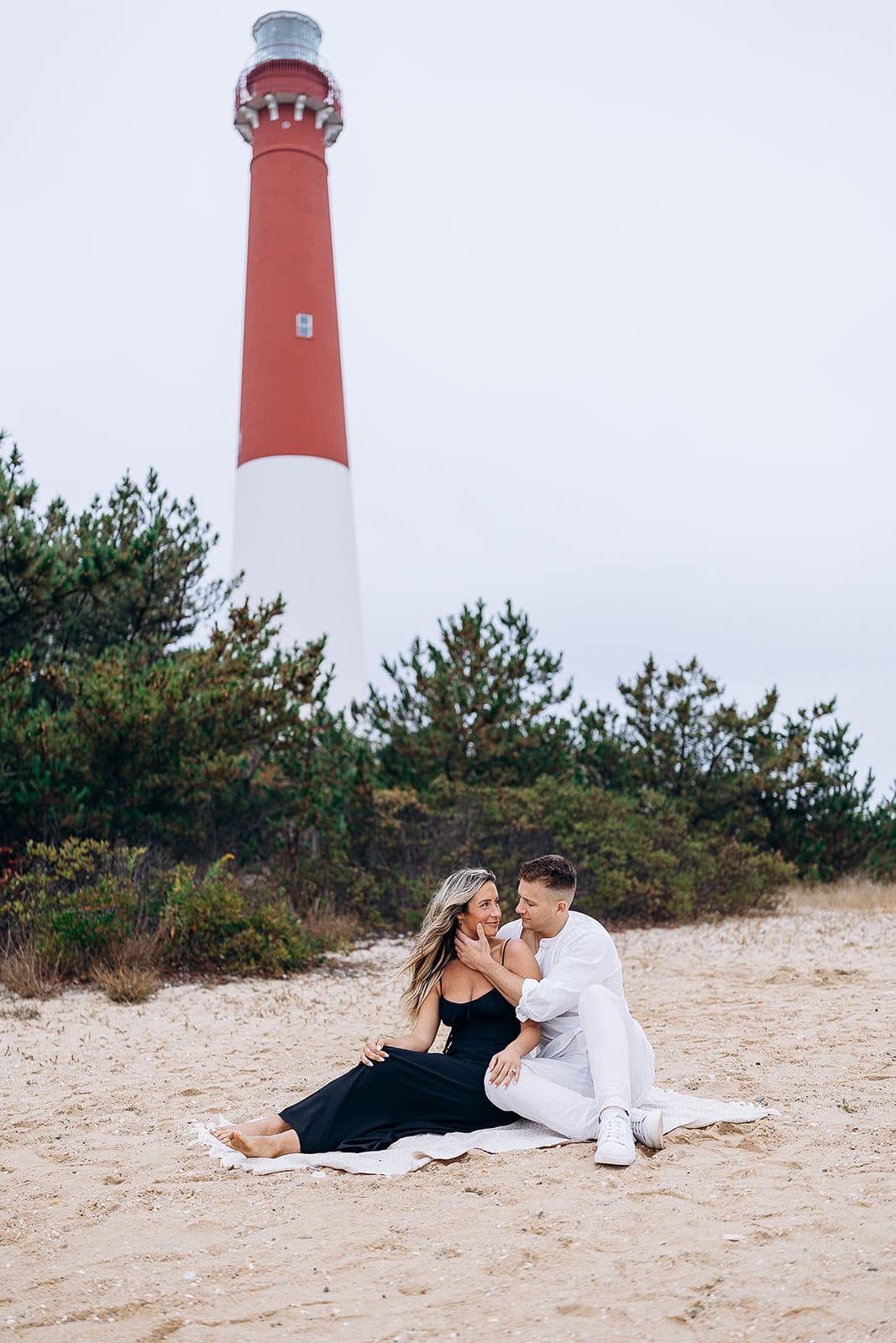 Couple seated on a sandy beach near Barnegat Light Lighthouse, surrounded by green shrubs under an overcast sky.