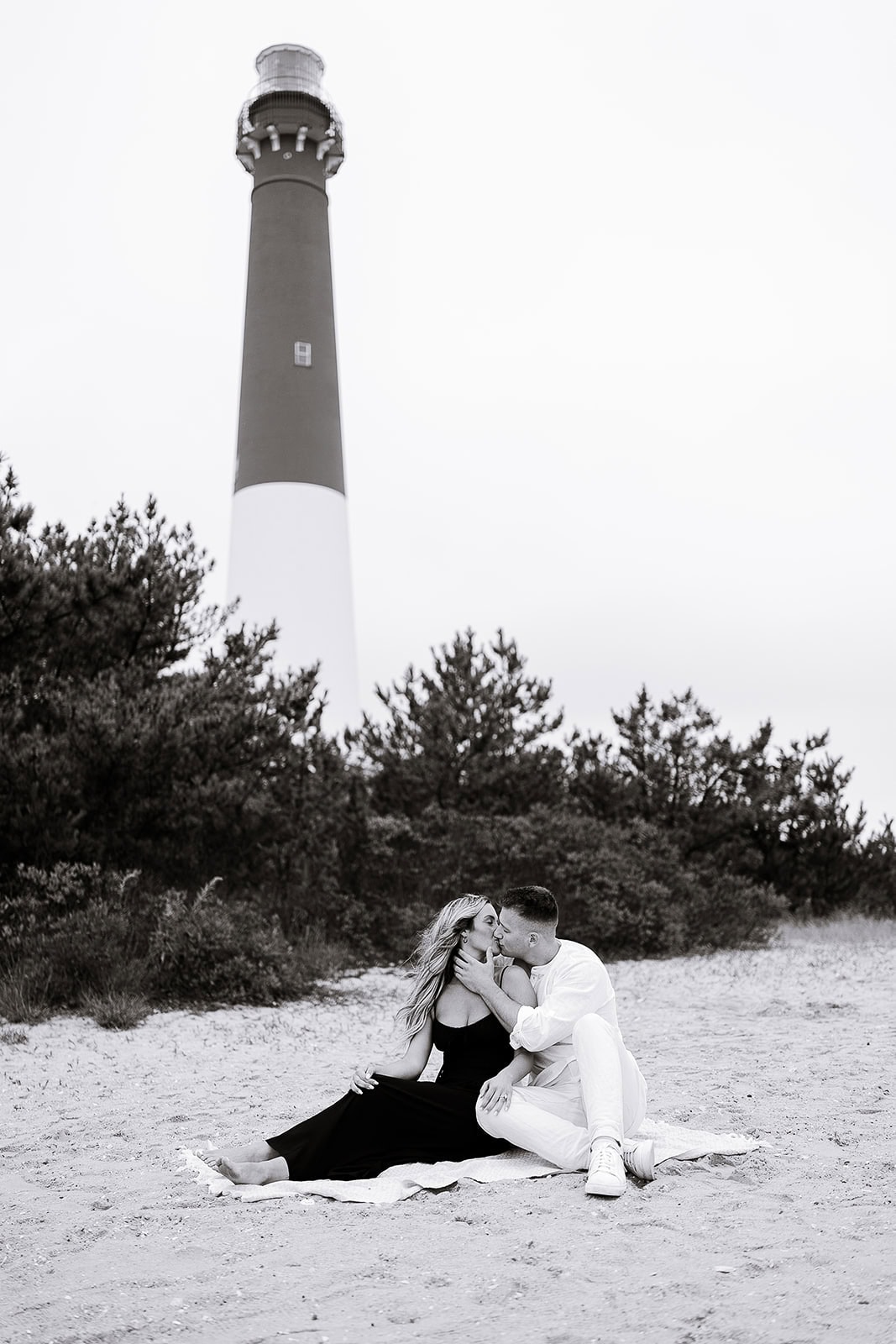 Couple sitting on sand kissing near Barnegat Light lighthouse with trees in the background, captured in black and white.