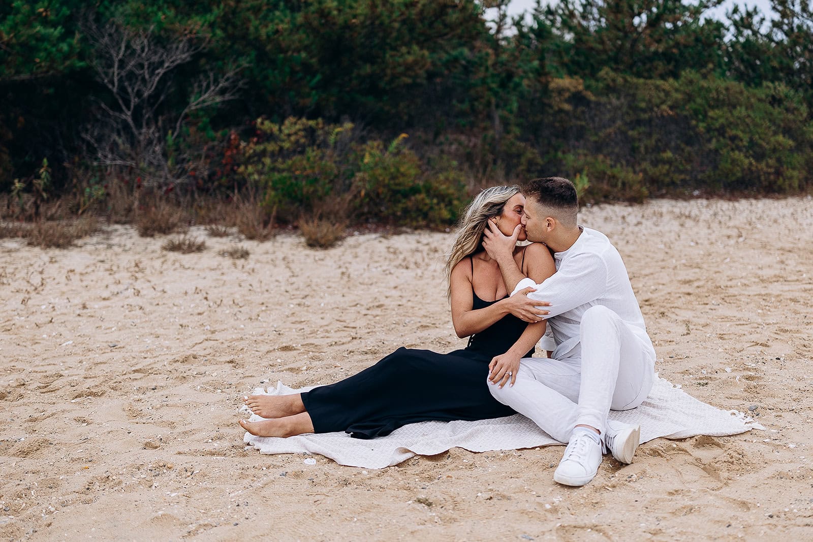 Couple sitting on a blanket on a sandy beach at Barnegat Light, NJ, sharing a tender kiss with natural greenery in the background.