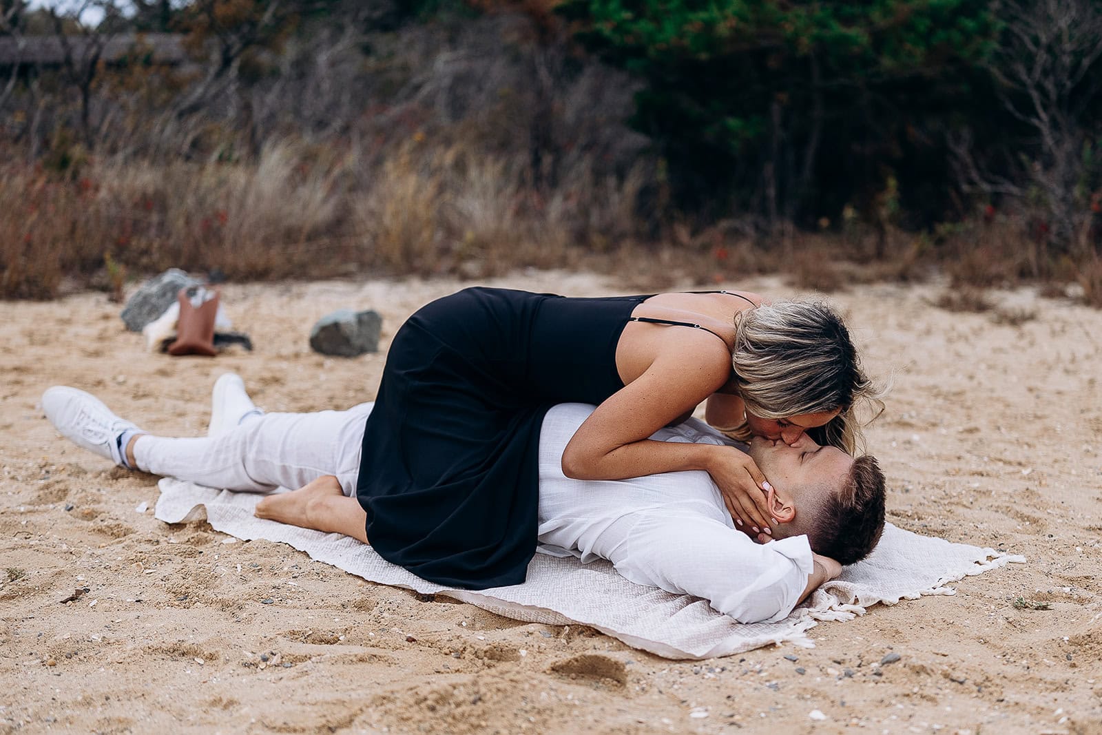 Couple lying on a sandy beach, woman in black dress kissing man in white shirt lying on a light blanket, surrounded by natural foliage.