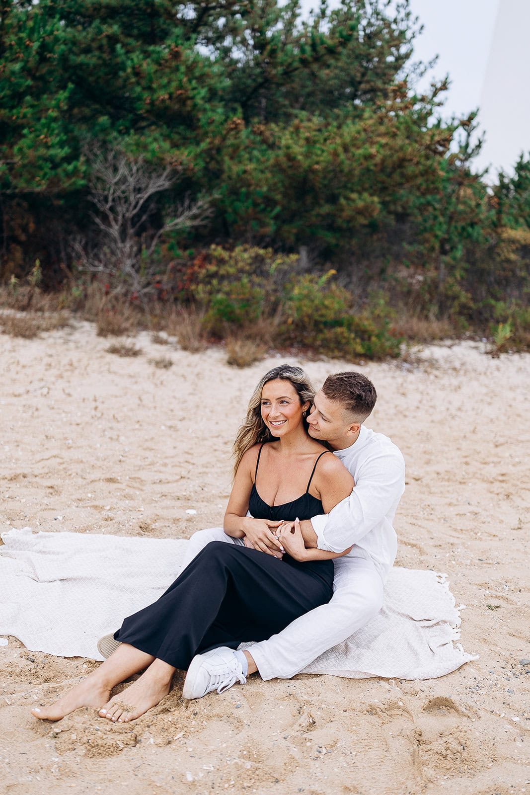 Couple seated on a light blanket on sandy beach with greenery in background, man in white shirt embracing woman in black dress.