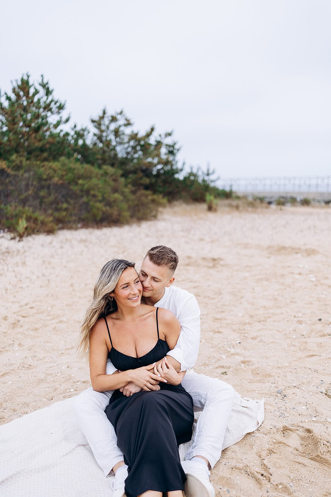 Couple seated on sandy beach with greenery in background, woman in black dress and man in white outfit embracing gently.