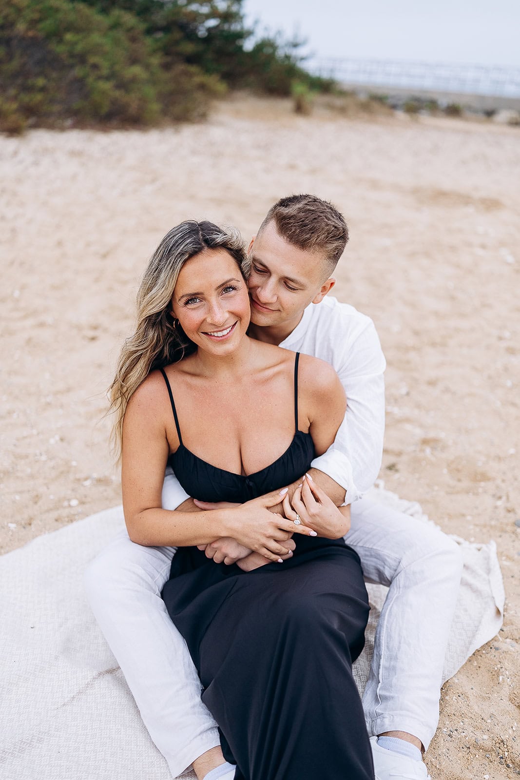 Couple seated on a sandy beach with the man in white embracing a woman in a black dress from behind, both smiling gently.