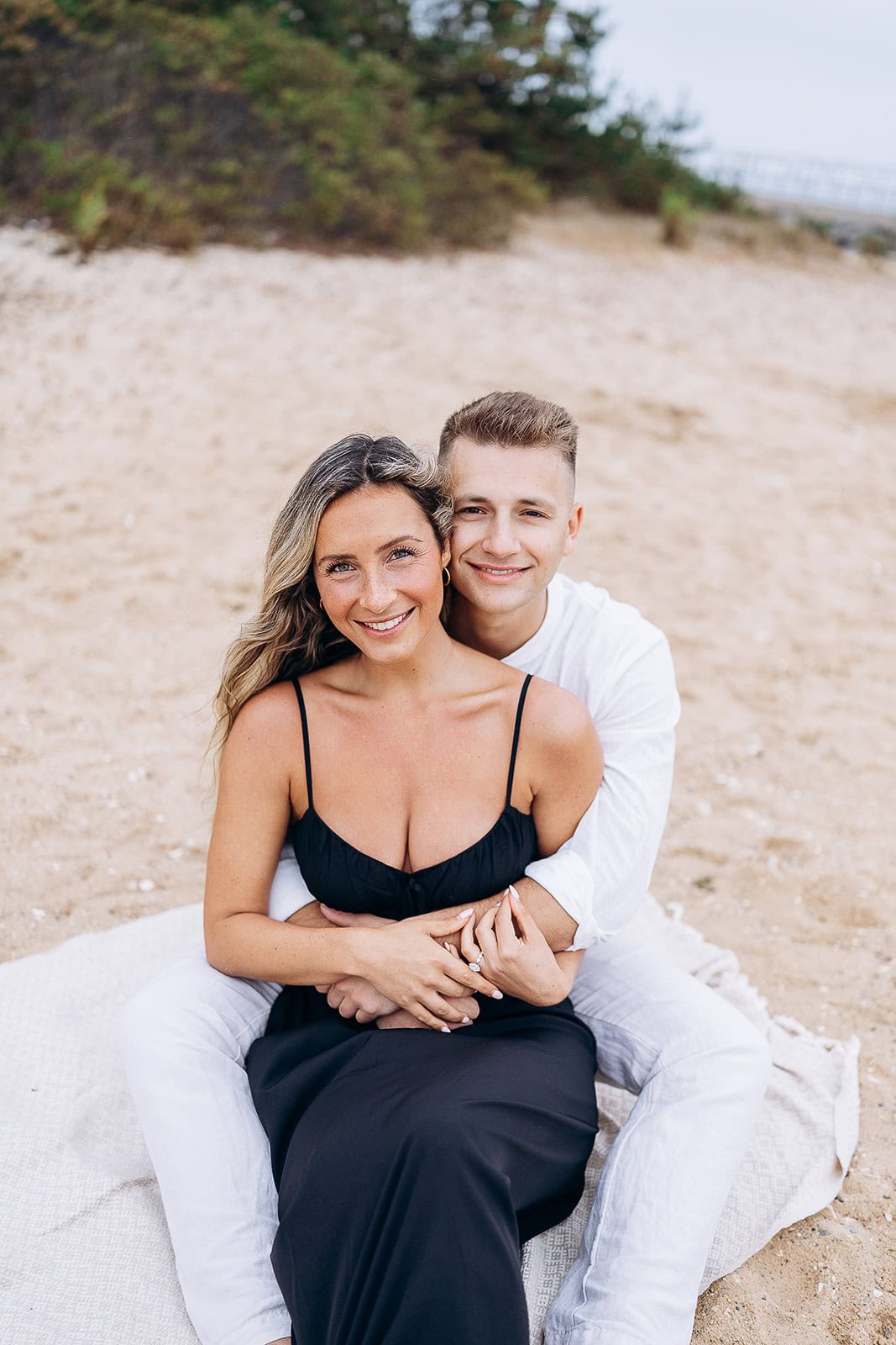 Couple seated on a blanket on sandy beach, woman in black dress, man in white shirt embracing from behind.