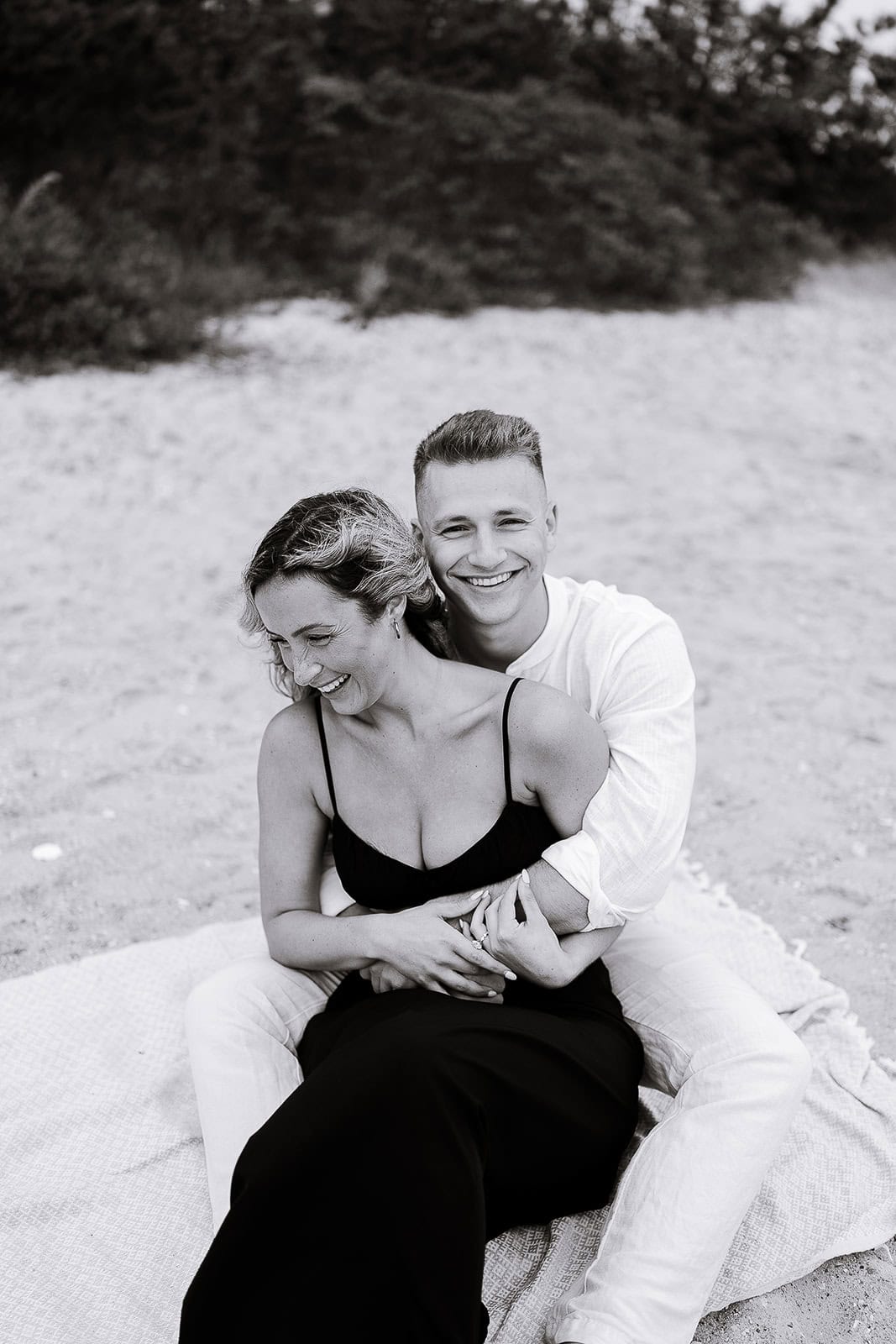 Black and white image of a couple seated on a blanket on the sand, the man embracing the woman from behind as they both smile, with blurred foliage in the background.