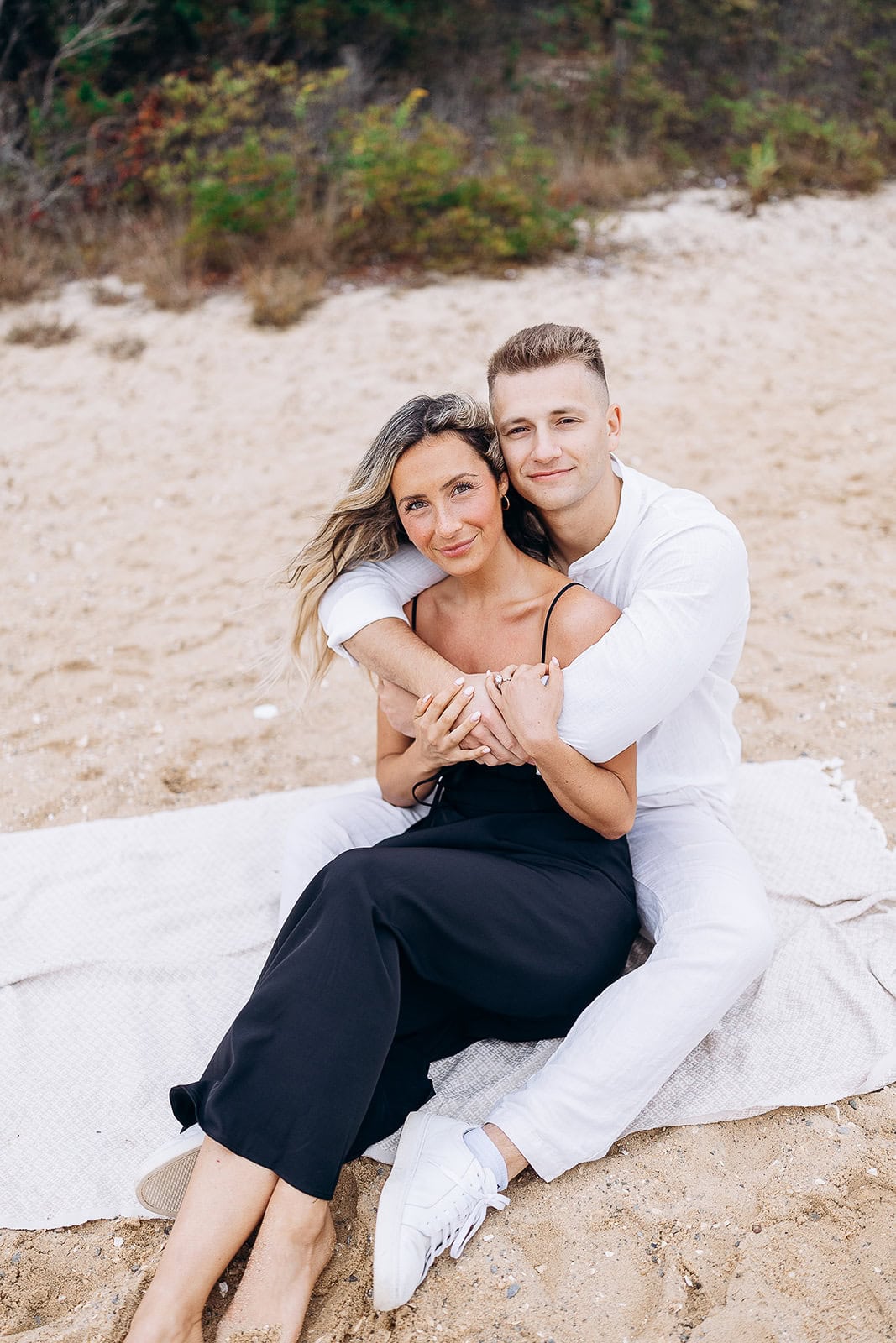 Couple seated on a blanket on sandy beach, man in white shirt embraces woman in black dress, natural greenery in background.