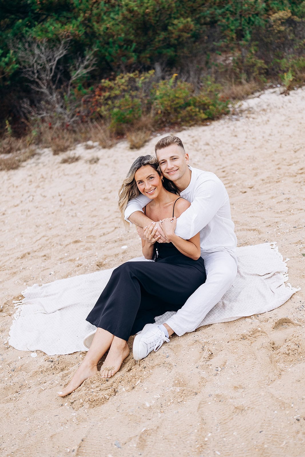Couple seated on a white textured blanket on sandy beach with greenery in background, embracing and smiling in Barnegat Light, NJ.