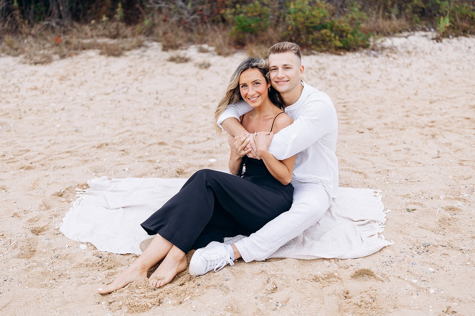 Couple sitting on a light blanket on sandy beach, man in white outfit hugging woman in black dress, natural foliage background.