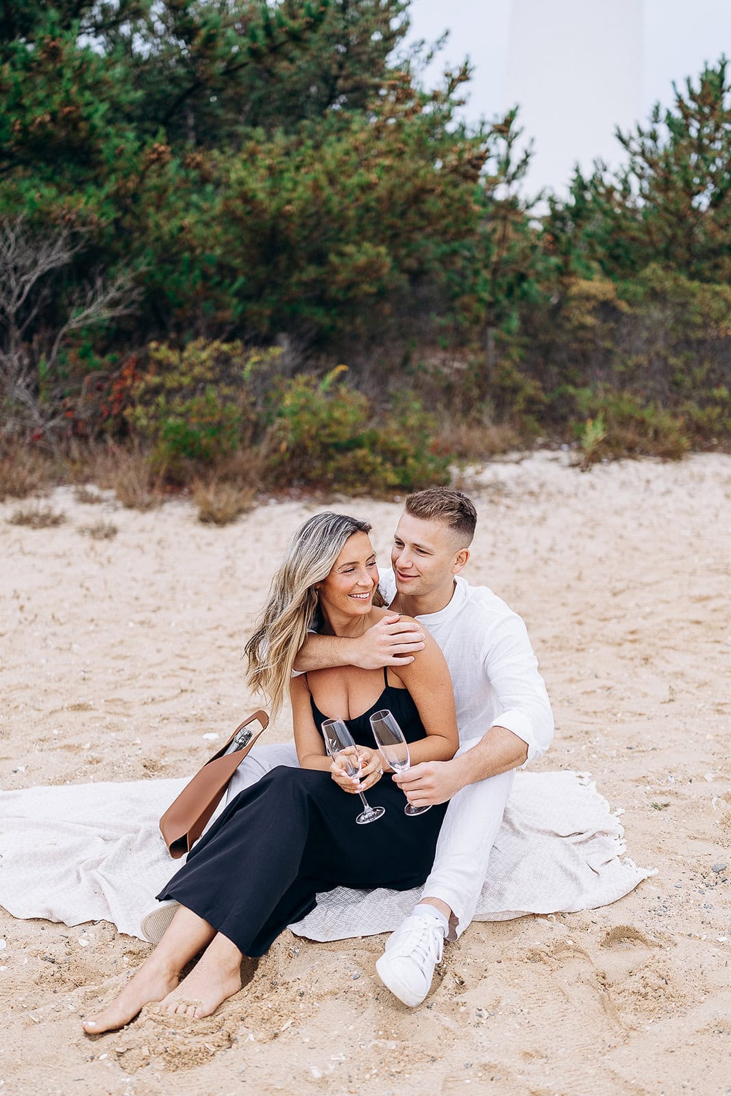Couple seated on a light blanket on sandy beach in Barnegat Light, woman in black dress holding champagne flutes, man in white outfit embracing her.