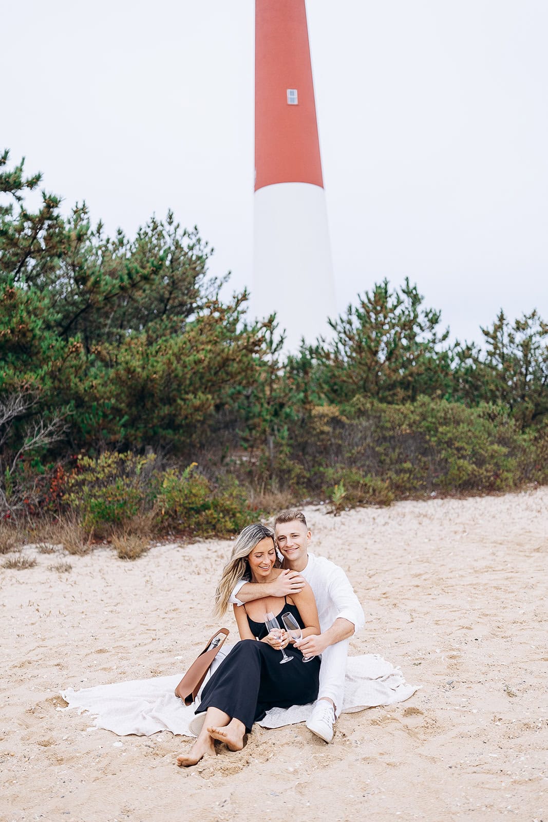Couple seated on sandy beach in front of Barnegat Light lighthouse, holding champagne glasses, surrounded by greenery.