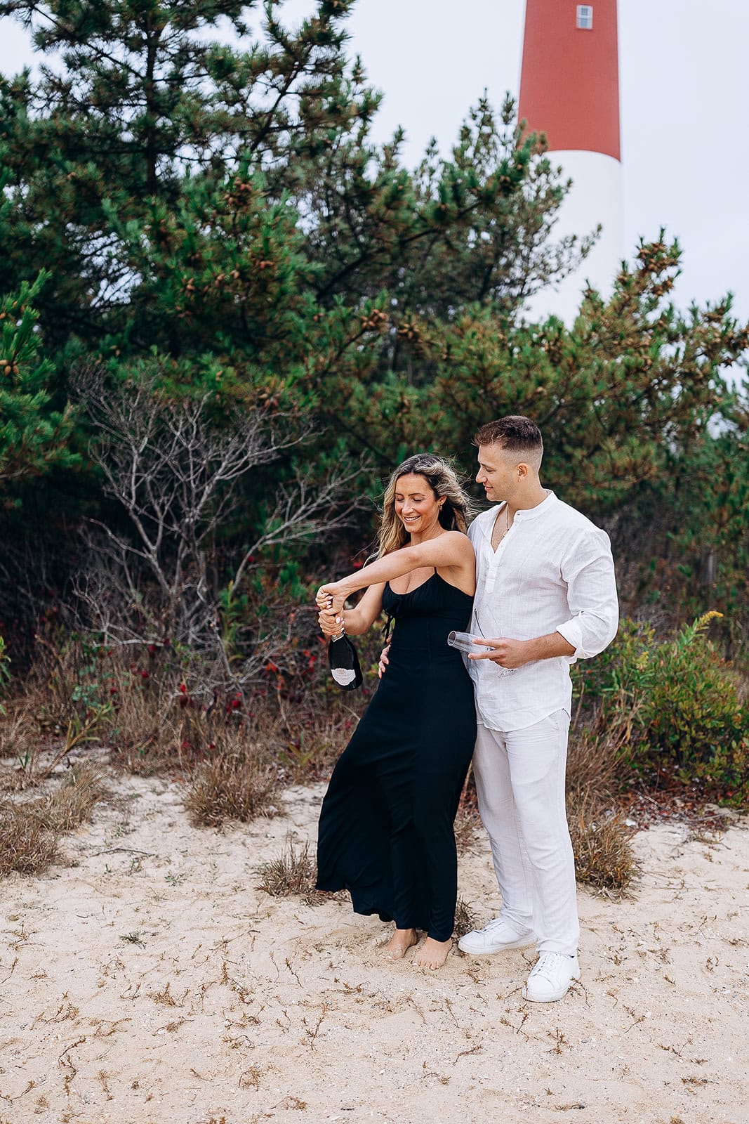 Couple on sandy beach at Barnegat Light, woman in black dress opening champagne bottle, man in white holding glasses, lighthouse and pine trees in background.