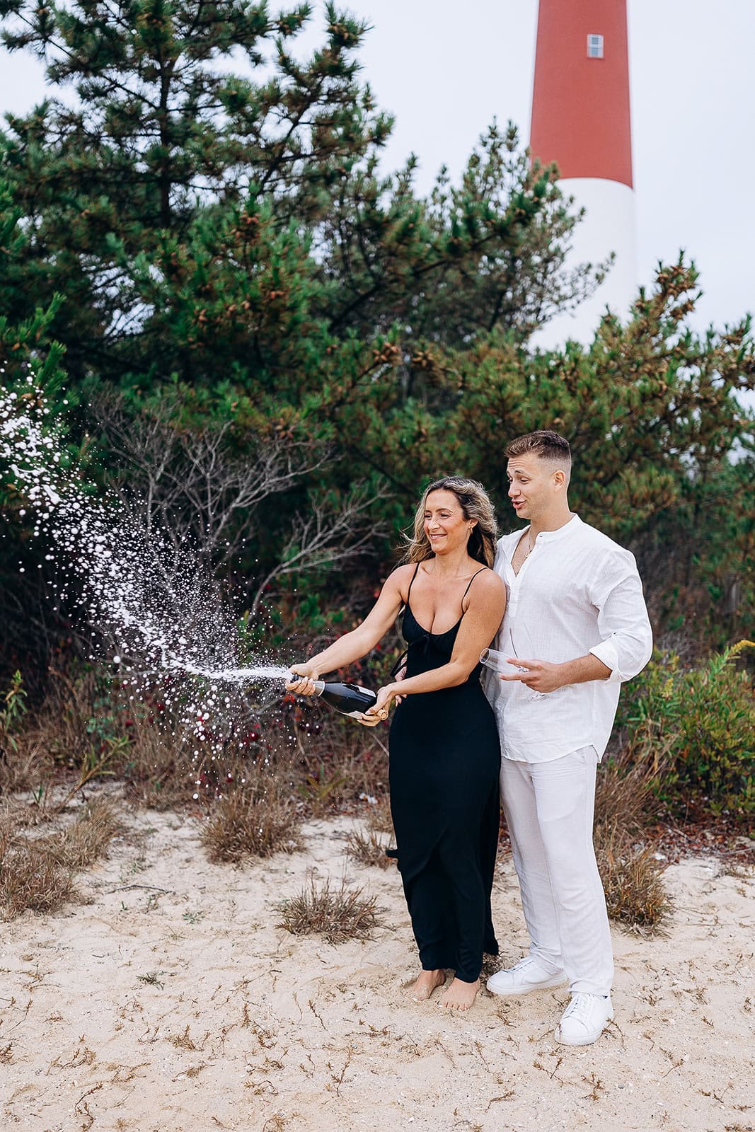 Couple on sandy beach near Barnegat Light lighthouse, woman in black dress popping champagne bottle, man in white outfit holding glass