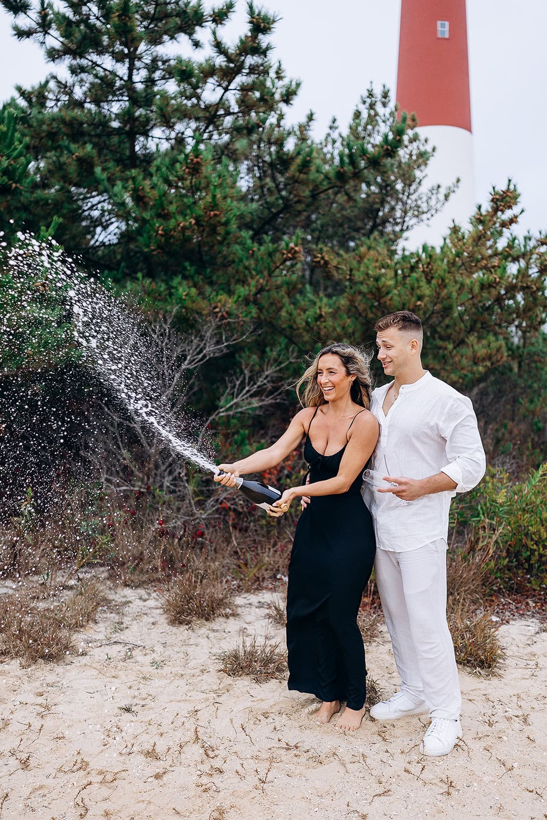 Couple on sandy beach at Barnegat Light, woman in black dress popping champagne bottle, man in white outfit holding glass, lighthouse and pine trees in background.