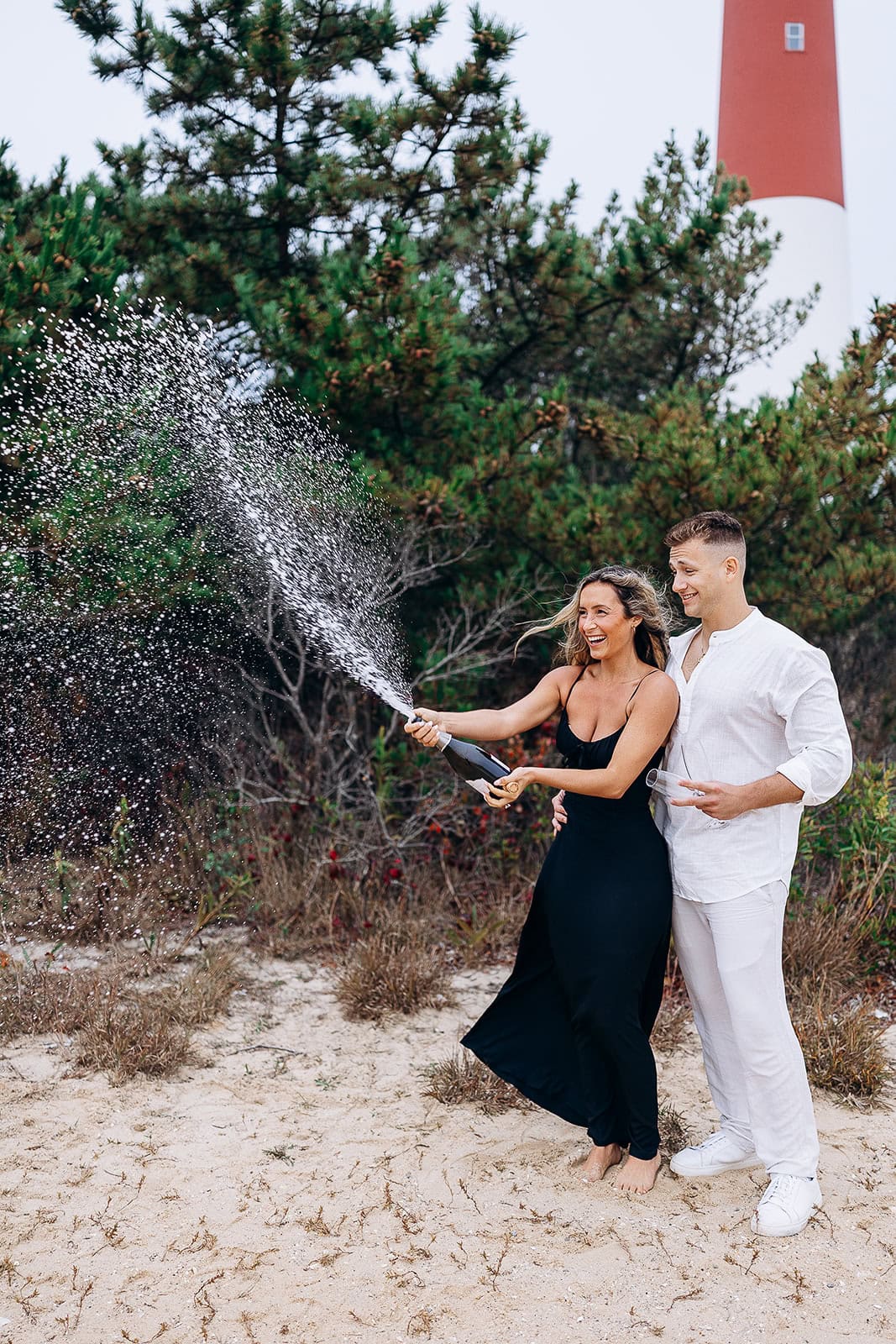 Couple on sandy beach at Barnegat Light, NJ, popping champagne with spray in front of pine trees and red and white lighthouse.