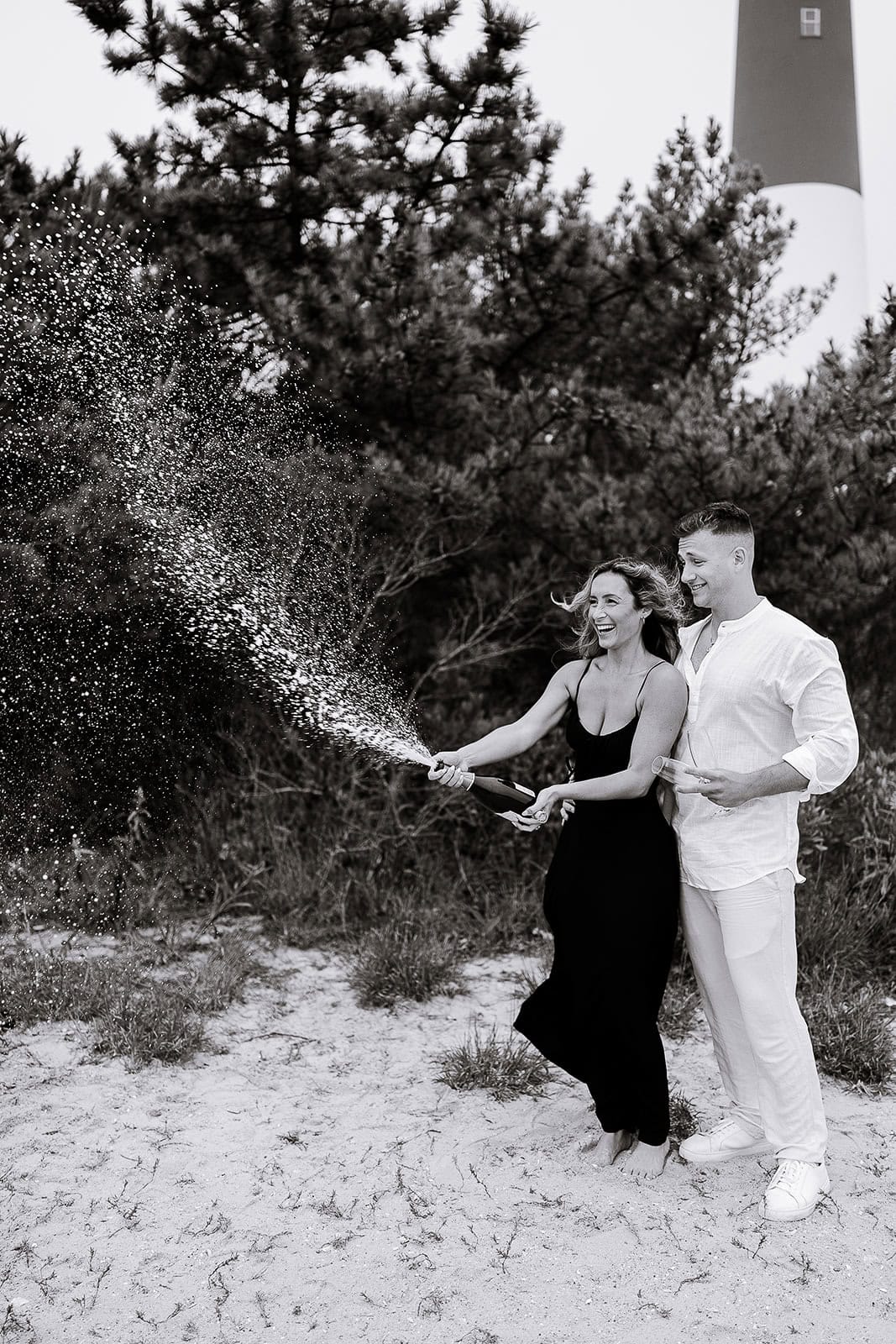 Couple celebrating on sandy ground at Barnegat Light, NJ, spraying champagne with trees and lighthouse in background.