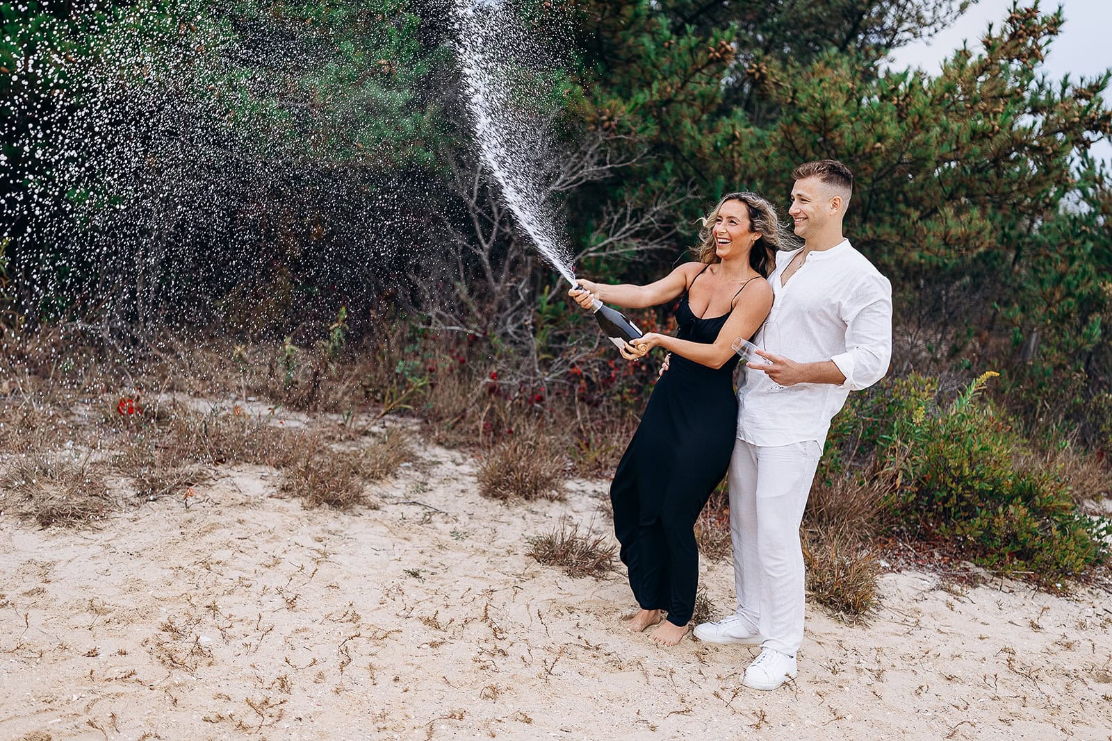 Couple on sandy beach with greenery in background, woman in black dress popping champagne bottle, man in white outfit holding glasses