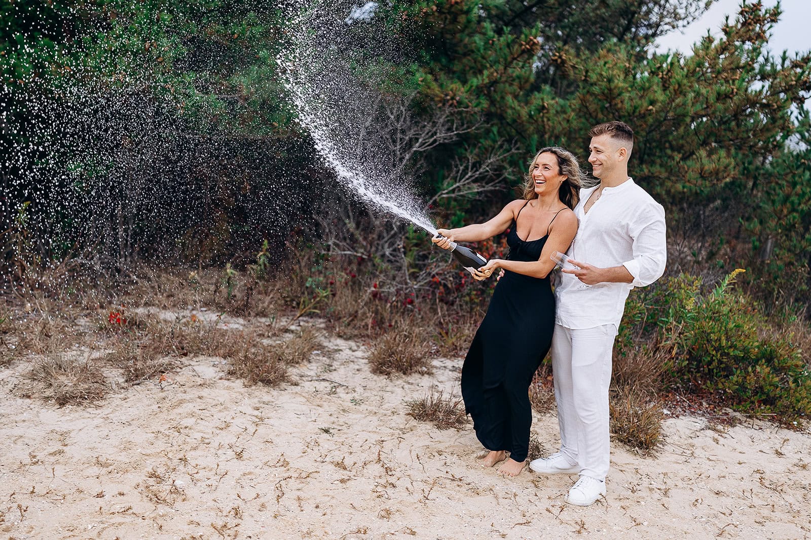 Couple on sandy beach surrounded by greenery, woman in black dress popping champagne with spray droplets, man in white outfit holding glasses.