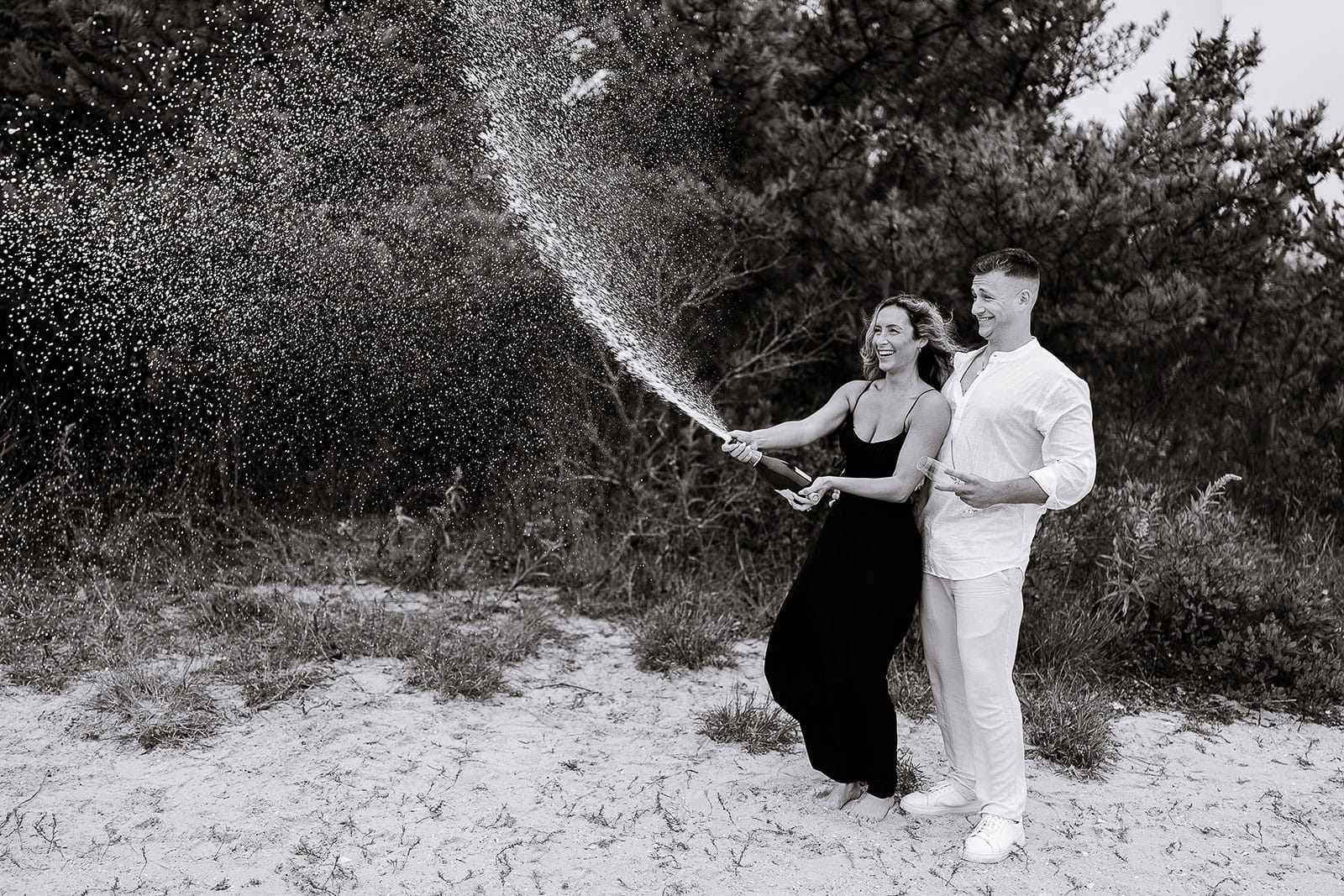Black and white image of a couple standing barefoot on sandy ground with shrubs behind them, the woman spraying champagne from a bottle while the man holds a glass, both smiling.